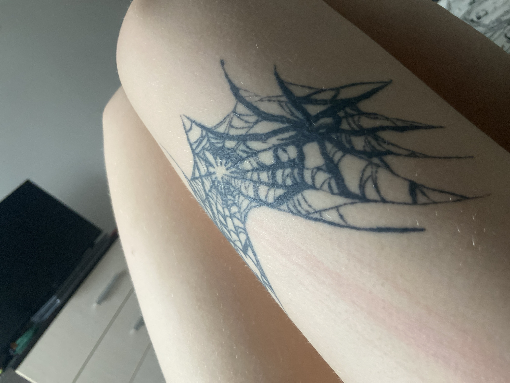 A close-up, eye-level, medium shot captures a tattoo of a spiderweb and spider on a person's leg. The tattoo is rendered in dark blue ink and is located on the upper thigh, near the knee. The leg is smooth and hairless, with a slight pinkish hue, possibly indicating recent application of the tattoo or skin irritation. The background is out of focus, but a portion of a white dresser with drawers and a dark rectangular object, possibly a television or monitor, can be seen. The lighting is soft and diffused, suggesting an indoor environment.