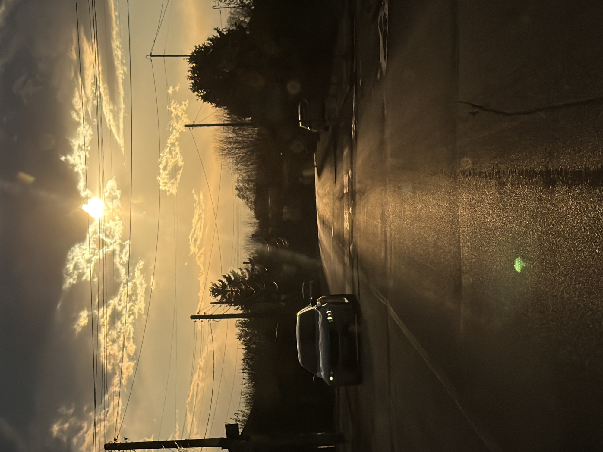 The image depicts a street scene, possibly in or near Yoakim Gruevo, Bulgaria. The sun is low in the sky, possibly during sunset or sunrise, casting a golden light. Clouds are visible in the sky, interspersed with telephone/electrical wires. The road is wet, reflecting the sunlight. There's a car parked on the right side of the road, facing the viewer. Another vehicle can be seen further down the road. Trees and bushes line the side of the road, silhouetted against the bright sky. Power poles are also visible along the road's edge.