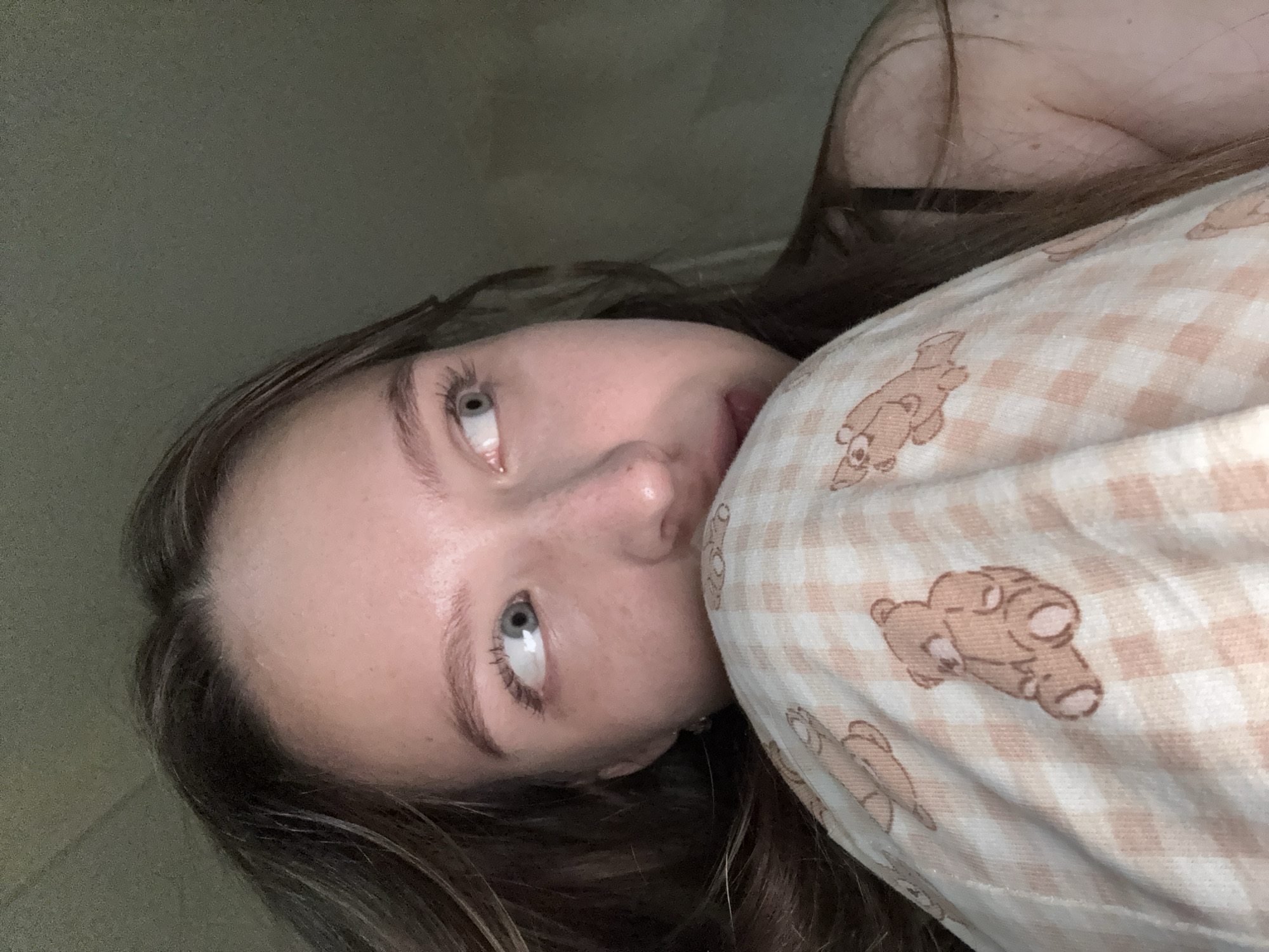 A young Caucasian female is depicted in a dimly lit indoor setting in the city of Chaikovsky, Russia. Her head is positioned to the left of the frame, with her body angled away from the viewer. She possesses long, dark brown hair, parted centrally, and light blue-grey eyes that are directed upwards and to her right, displaying a wide-eyed gaze. Visible freckling is present on her nose and cheek. Her lower face, from the nose down, is partially obscured by a light-colored fabric. This fabric features a pale peach and white gingham check pattern adorned with stylized brown cartoon bears in a sitting posture, alongside less distinct, elongated brown shapes that resemble fish. This material appears to be clothing or bedding. A dark strap, consistent with an undergarment or top, is visible over her right shoulder. The background is a uniform, dark, out-of-focus grey-green, indicating low ambient light conditions.