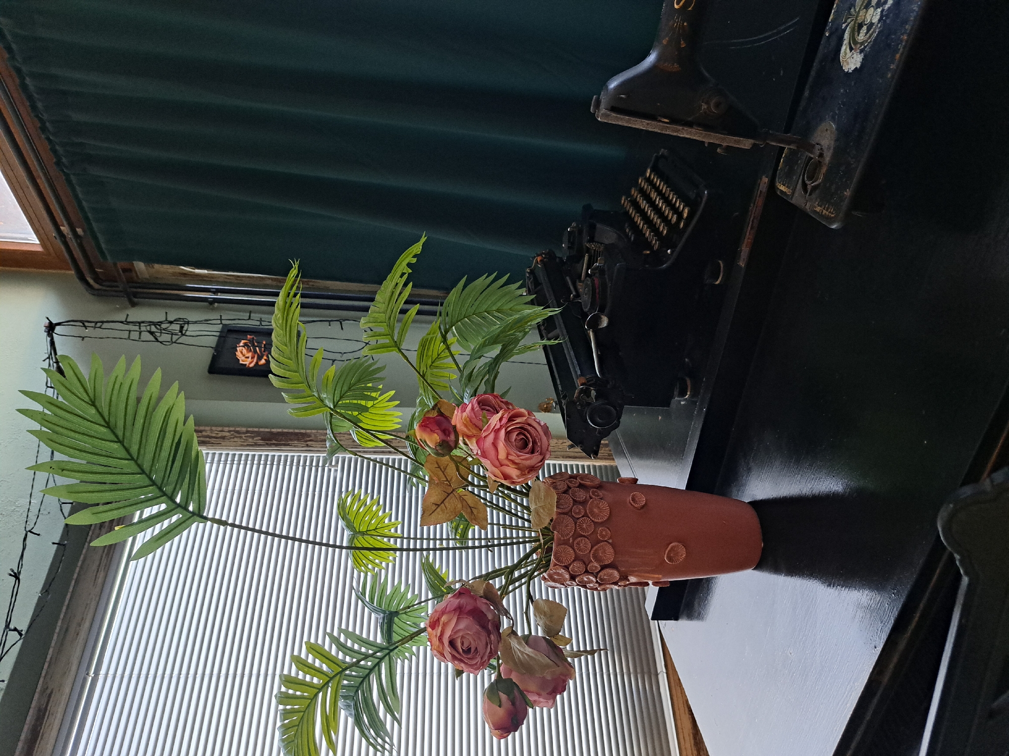 In an indoor setting, possibly a room in a house, a black antique typewriter is placed on a dark, polished table. To the right of the typewriter, a part of an antique sewing machine is visible. In the foreground to the left, a pink vase filled with artificial flowers and palm leaves stands on the table. Behind the objects, a window with horizontal blinds is visible, and a dark green curtain hangs on the right side of the frame. The overall scene suggests a collection of vintage items displayed in a domestic environment.