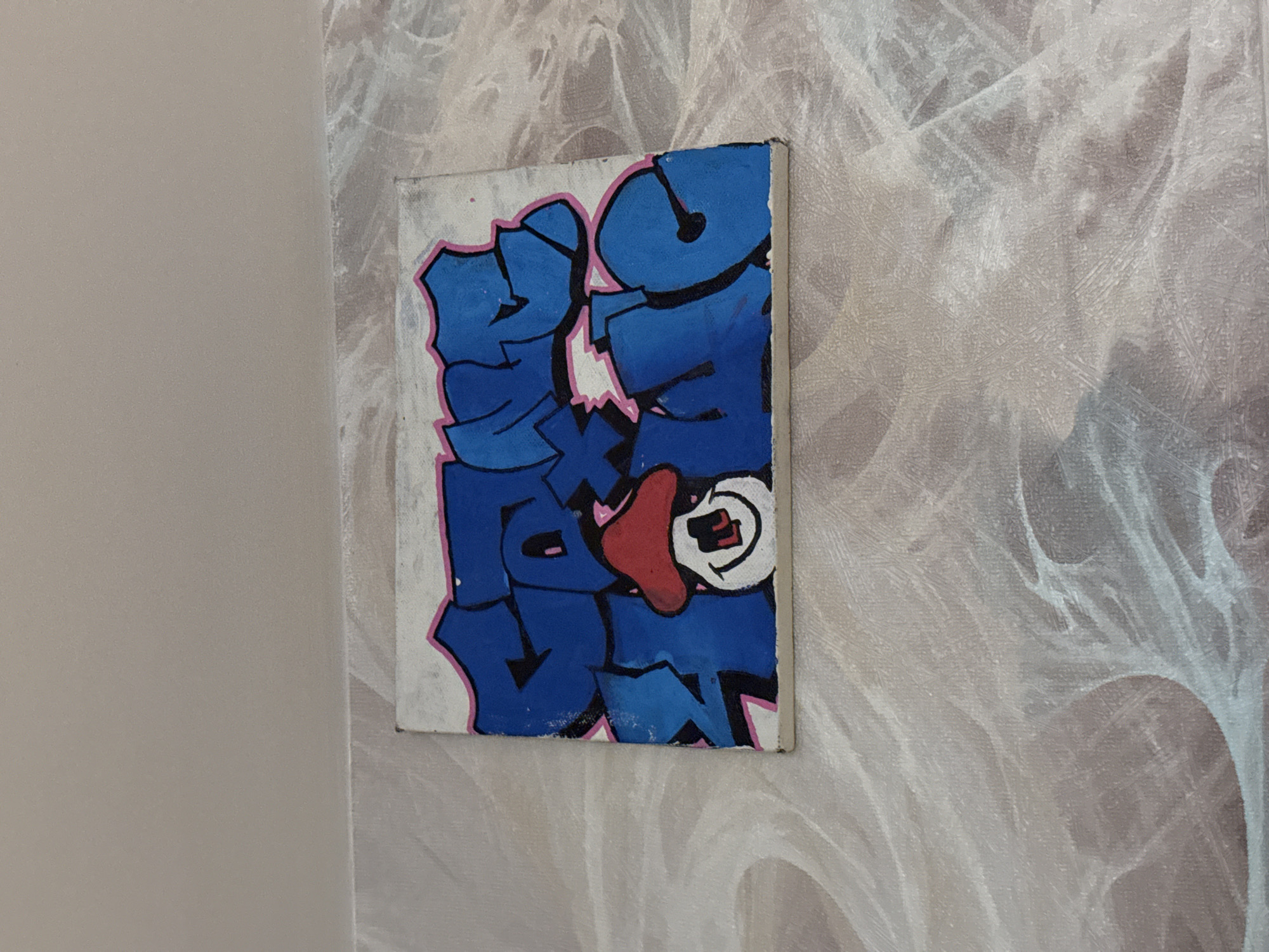 The image shows a close-up, eye-level shot of a graffiti art piece on a canvas. The graffiti features bold blue bubble letters outlined in pink and black, with a stylized cartoon face with a red hat and white features incorporated into the lettering. The canvas is placed against a textured, light-colored wall, possibly wallpaper, with a light and shadow pattern. The top of the image shows a plain, light-colored ceiling. There is no indication of the specific location in Saint Petersburg, Russia, as the image focuses solely on the artwork and its immediate backdrop.