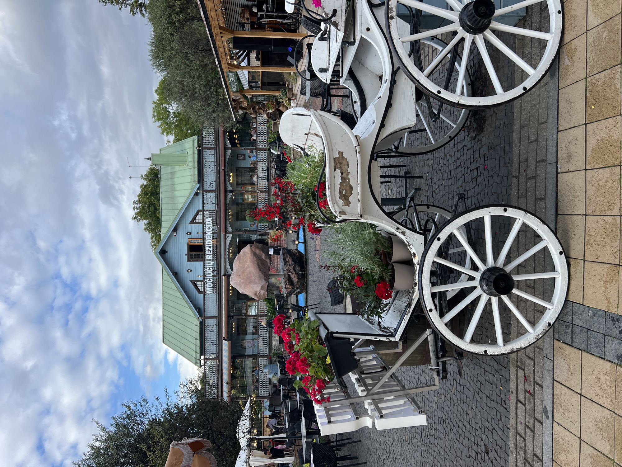 The image shows a white horse-drawn carriage in the foreground, parked on a cobblestone street. The carriage has large spoked wheels and is decorated with red flowers in pots. Behind the carriage, there is a blue building with a green roof, which appears to be a restaurant or cafe with outdoor seating. The building has a sign that reads "MOCIUCIU REZIDENCIJA" in large letters. There are tables and chairs set up outside the building, and several people can be seen dining. To the right of the building, there is a covered outdoor area with more tables and chairs, and a stage area with musical equipment. The overall scene suggests a lively outdoor dining and entertainment area in the city of Palanga, Lithuania. The sky is cloudy, with patches of blue visible.