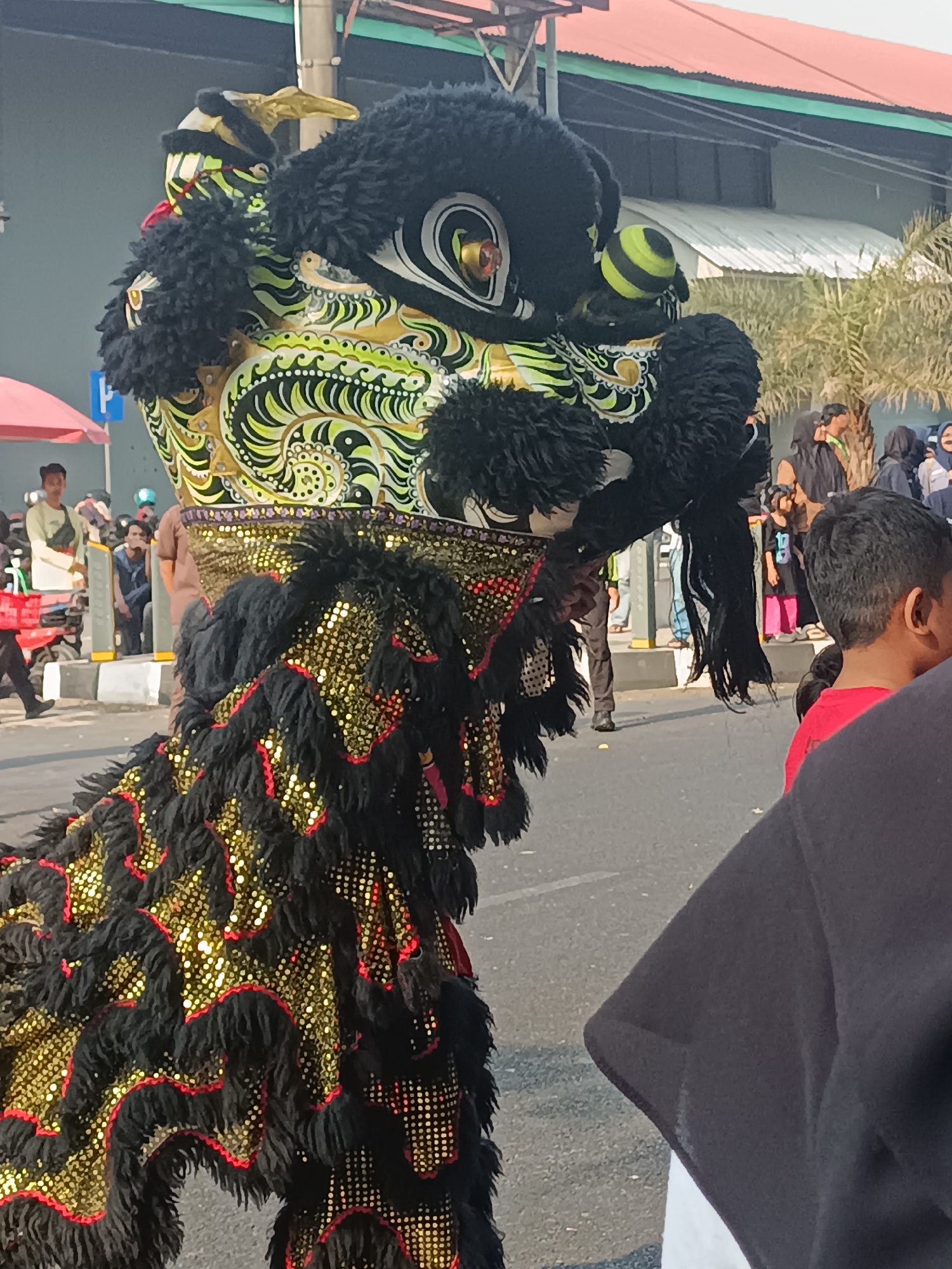 In Sleman, Indonesia, a colorful lion dance performance is underway on a street. The focal point is the head and part of the body of a lion dance costume. The head is elaborately decorated with black fur, green and yellow swirling patterns, and large, expressive eyes with red and yellow accents. A spherical, yellow and black striped object protrudes from its forehead. The body of the lion is covered in black fur with gold sequins arranged in a wavy pattern, bordered by red fringing. A person in a red shirt is visible in the foreground, with their back to the camera, obscuring the lower part of the lion's body. In the background, several people are observing the performance. Behind them, palm trees and buildings with red and green roofs are visible, suggesting an outdoor urban setting. The street is paved, with a curb and what appears to be a sidewalk or pedestrian area. The lighting indicates it is daytime, with shadows cast on the ground.