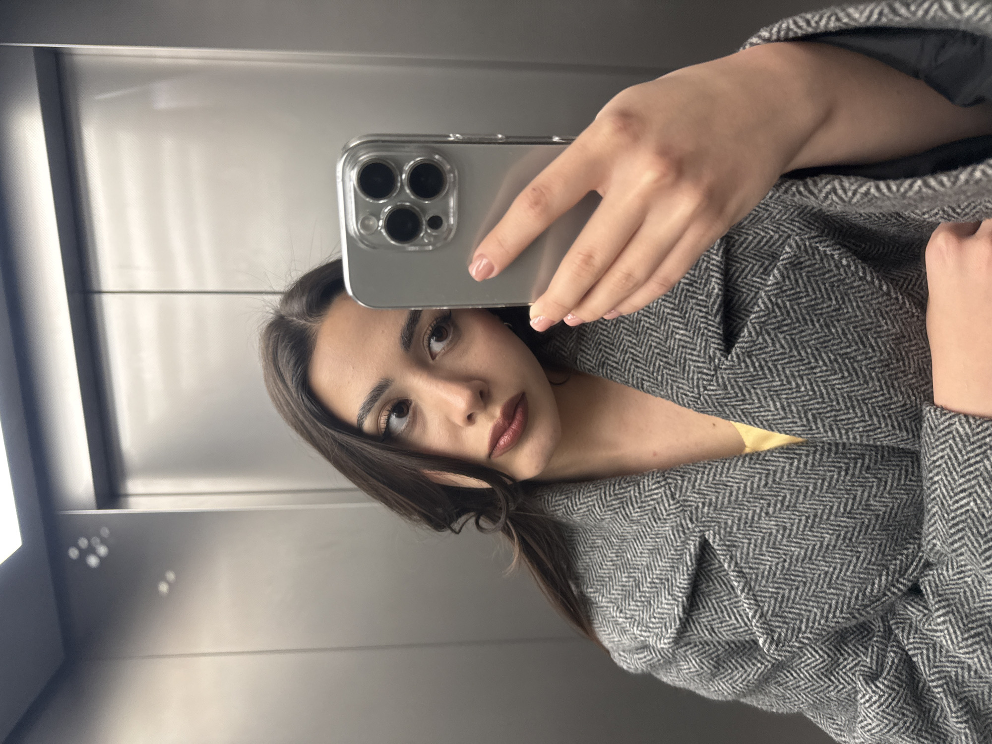 A young woman takes a selfie in an elevator with a modern smartphone. She is holding the phone in her right hand, and the back of the phone is visible, featuring three camera lenses. The woman has dark hair and is wearing a grey herringbone patterned coat. Behind her, the elevator interior consists of plain metal panels. The location is unknown and cannot be determined from the image.