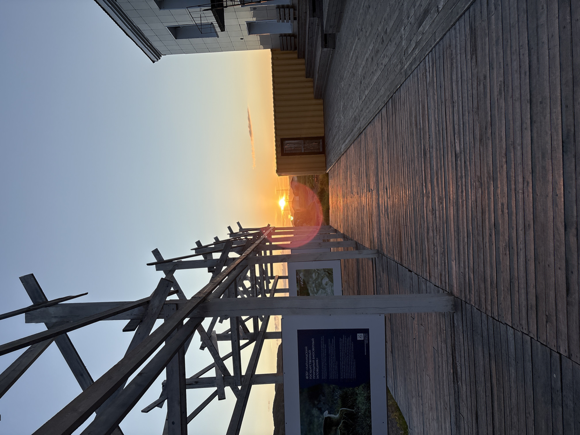 The image depicts an outdoor scene at sunset in Teriberka, Russia. In the foreground, a wooden boardwalk stretches into the distance. To the left, a wooden structure with exposed beams is visible, supporting informational panels with text and images. To the right, a modern building with white siding and a prominent staircase leads up to an entrance. A yellow shipping container is positioned next to the building. The sun is low on the horizon, casting a warm glow across the scene and creating lens flare. The sky is clear with a gradient of colors from pale blue to orange. The environment appears to be coastal or open terrain, with distant hills or landforms visible against the sunset. There are no people present in the image.