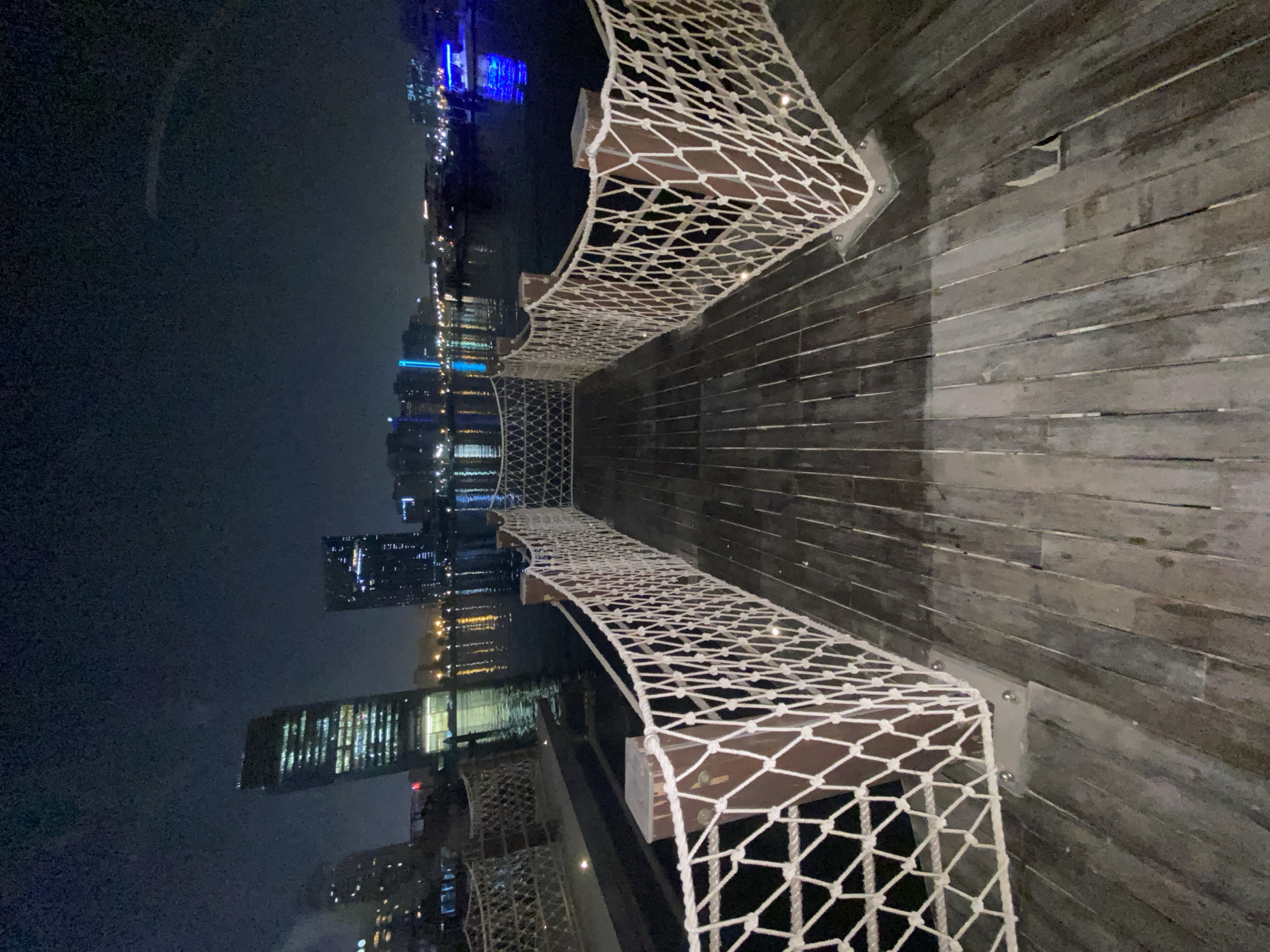 The image captures a night scene of a wooden pier in Deira, United Arab Emirates. The pier features railings made of thick white rope woven into a net-like pattern, supported by wooden posts. Beyond the pier, a body of water reflects the lights of the city skyline, which is illuminated by tall buildings and the lights from boats. One boat in the foreground, on the right side of the image, has bright blue lights. The sky is dark, indicating nighttime. The wooden planks of the pier are visible in the foreground, showing signs of wear.