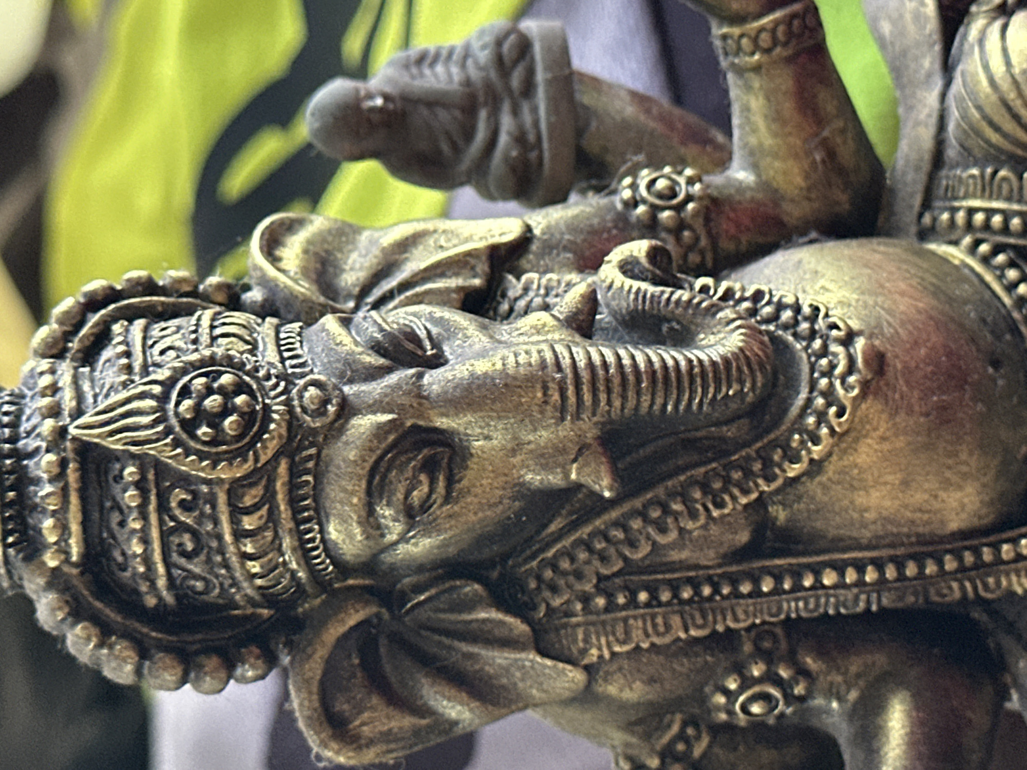 The image is a close-up, eye-level, medium shot of a bronze statue of the Hindu deity Ganesha. The statue is ornate, with intricate carvings on its headdress, jewelry, and body. Ganesha's trunk is curled to the left, and his eyes are closed. To the right of Ganesha, a smaller, dark-colored figurine of a meditating person is visible, also appearing to be made of bronze. The background is out of focus and features a bright green fabric with a black pattern. The statue's metallic surface shows signs of wear and a patina that suggests age. The scene appears to be indoors or in a sheltered outdoor area, possibly a shop or a home in Karon, Thailand, given the mention of Karon, Thailand, in the prompt, but there is no definitive visual evidence of this specific location.