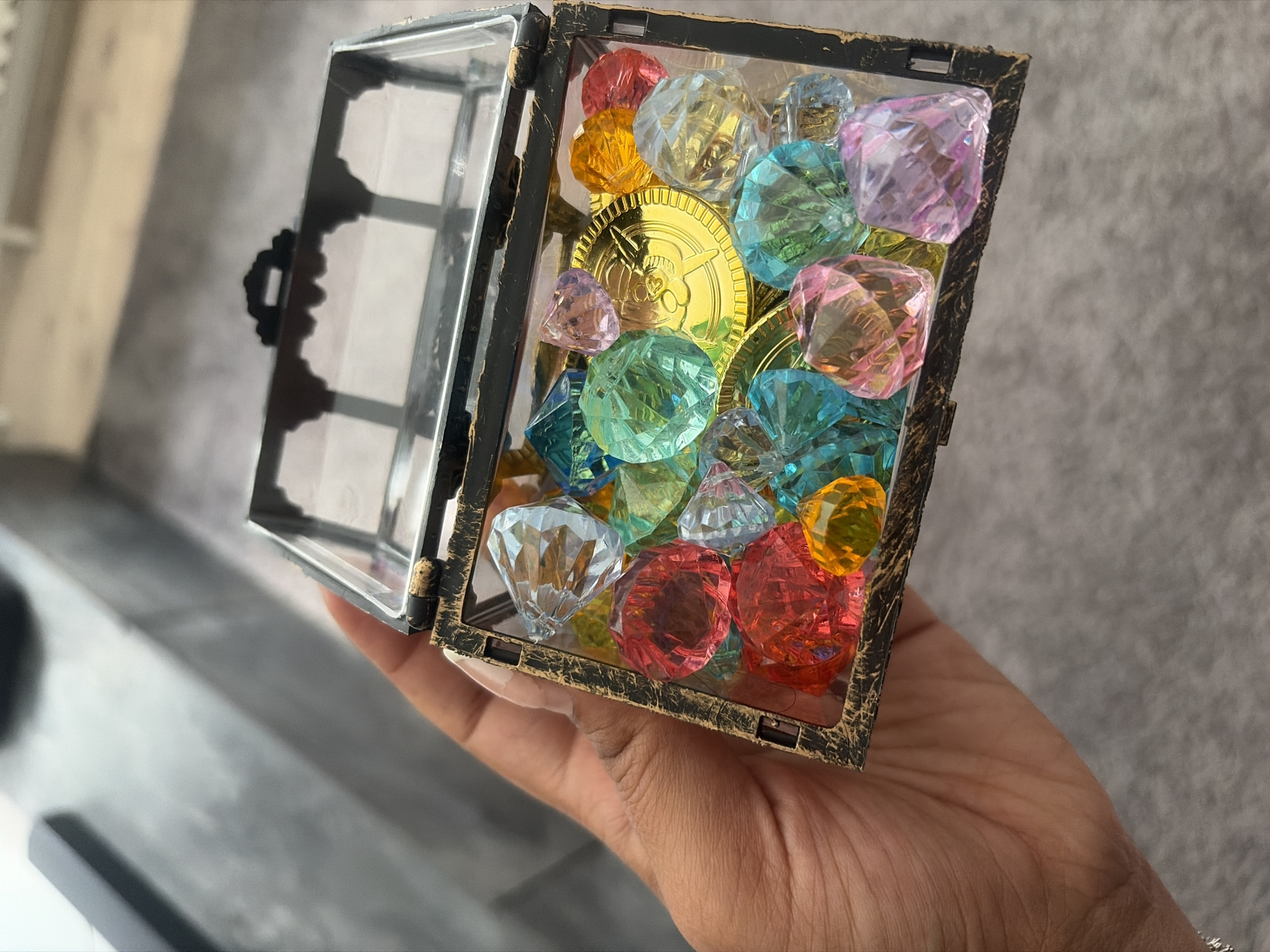 A close-up, high-angle shot shows a person's hand holding a small, open treasure chest filled with colorful, faceted acrylic gems and several gold-colored plastic coins. The chest has a distressed, antique gold finish on its frame and a clear glass top. The gems are various colors, including red, orange, yellow, green, blue, and pink, and are cut into geometric shapes. The coins appear to be play money with a raised design. The background is blurred and appears to be an indoor setting with gray carpeting and a darker surface in the upper left. There are no discernible landmarks or clues to indicate a specific location in Duisburg, Germany.