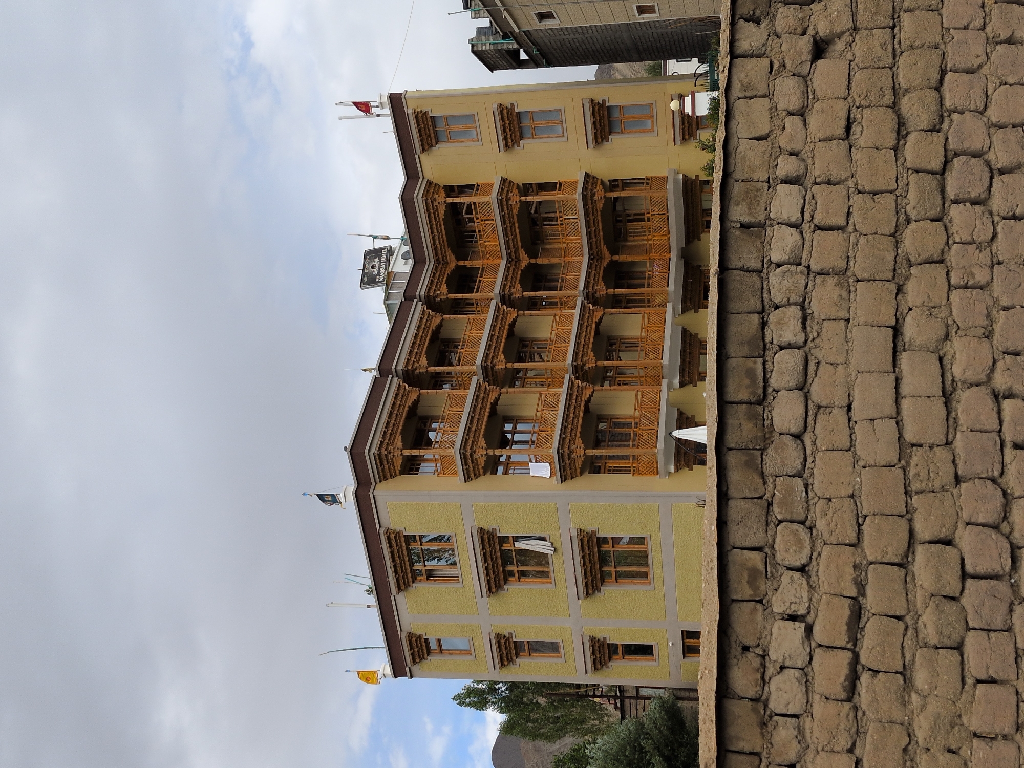 The image shows a multi-story building in Leh, India, with traditional Ladakhi architecture. The building is painted a pale yellow with brown trim and features ornate wooden balconies on multiple levels. There are several windows on each floor, some with white curtains visible. On the roof, there are flags and a sign for the "IMBANSAY HOTEL". The building is situated behind a rough, stone wall constructed of uneven, square-shaped bricks. In the background, there are trees and a rocky mountain. The sky is overcast with clouds.