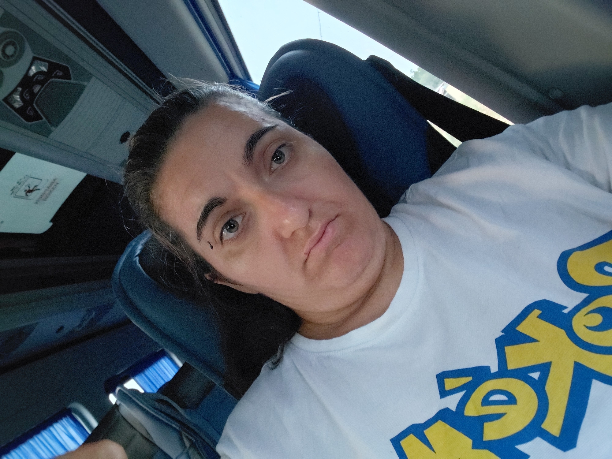 A woman with dark hair is taking a selfie from her seat on a bus. She is wearing a white t-shirt with a blue and yellow graphic design. The bus interior is visible, with blue seats and windows. A safety sign is mounted above the seats, and part of a control panel is visible. The scene suggests the woman is traveling.