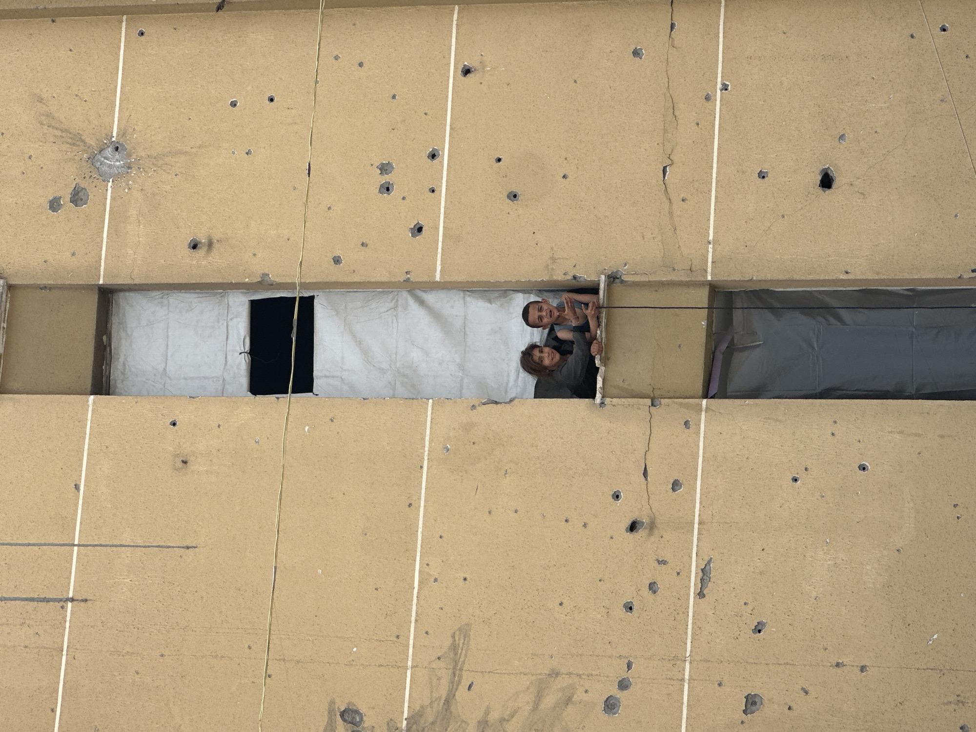 The image shows a portion of a building facade with numerous holes, some larger and more pronounced than others, indicative of potential damage from projectiles. The facade is divided by vertical white lines. A rectangular opening in the building reveals two individuals, one appearing to be a young male, looking out. One section of the opening is white, another black, and a third section has gray plastic sheeting. The environment seems to be urban. Specific city locations are not discernible from the image.