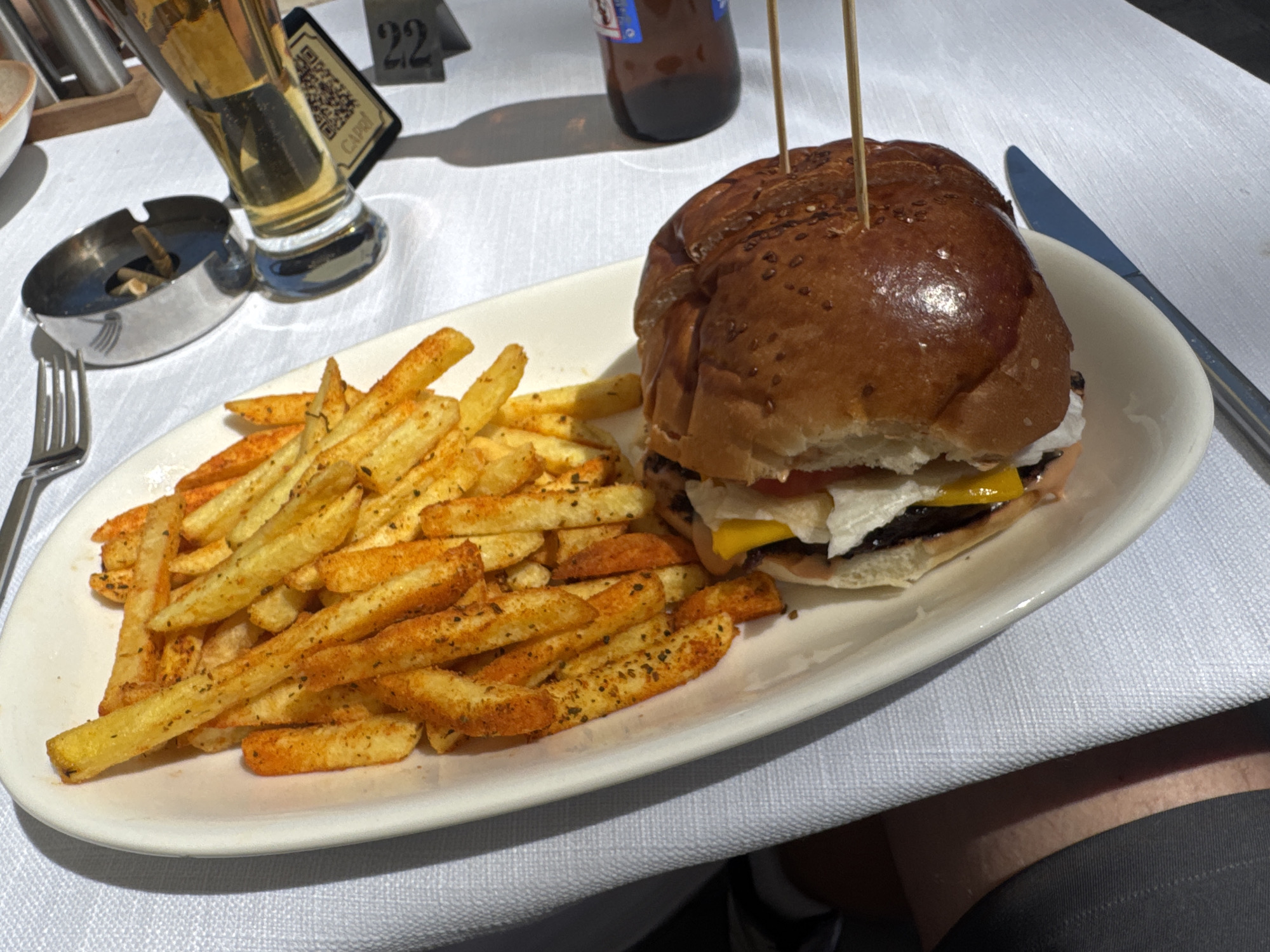 An overhead medium shot captures a white oval plate filled with a large portion of seasoned french fries and a gourmet burger on a white tablecloth. The burger, skewered with two wooden toothpicks, is topped with a glossy, sesame-seed-adorned bun. Beneath the bun, layers of lettuce, tomato, cheese, and a dark patty are visible. The french fries, dusted with a reddish-orange seasoning and flecks of green herbs, are piled to the left of the burger, some overlapping. To the left of the plate, a fork rests near a metal ashtray containing several cigarette butts and an overflowing glass of amber-colored liquid, likely beer. Behind the glass, a black QR code stands upright. In the background, a table number, "22," is visible. The scene suggests a dining setting, likely outdoors given the natural light illuminating the food and tablecloth. The table number implies a restaurant or cafe. The presence of the ashtray with cigarette butts and the QR code suggests a casual dining establishment, possibly in the Prince Islands, Turkey, where such amenities are common.