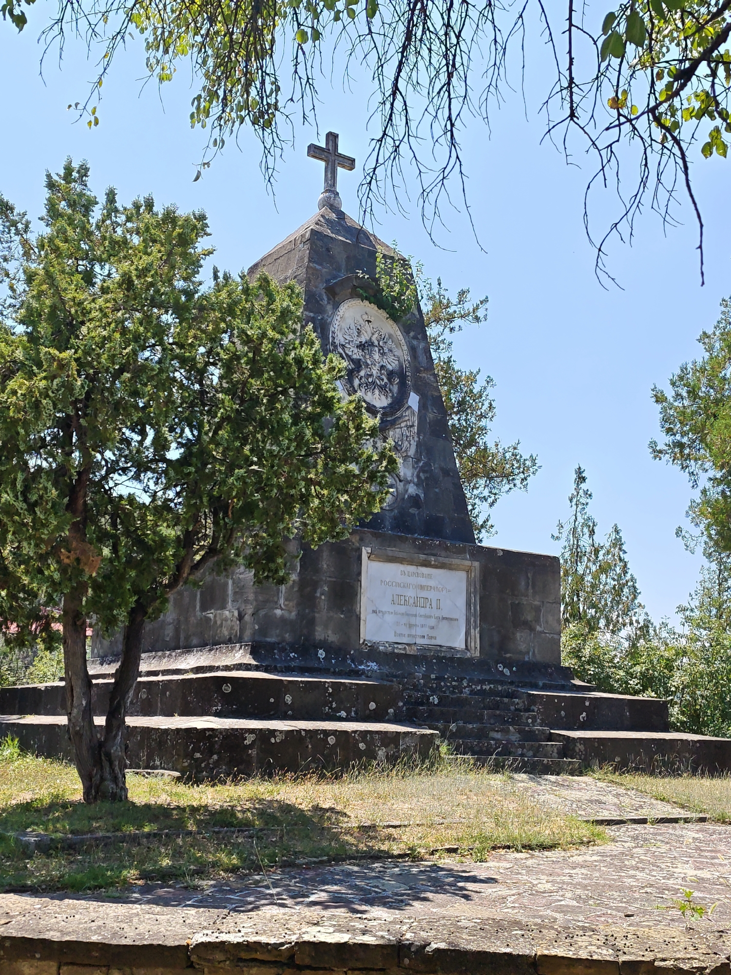 The image depicts a monument located in the city of Lovech, Bulgaria. The monument is a dark stone pyramid with a cross on top, and features a bas-relief circular emblem on one side and a plaque with inscription on another. A conifer tree is situated to the left of the monument, and stairs lead up to its base. The surrounding environment consists of grass, trees, and a clear blue sky, suggesting an outdoor setting. The overall scene is serene and appears to be a historical site.