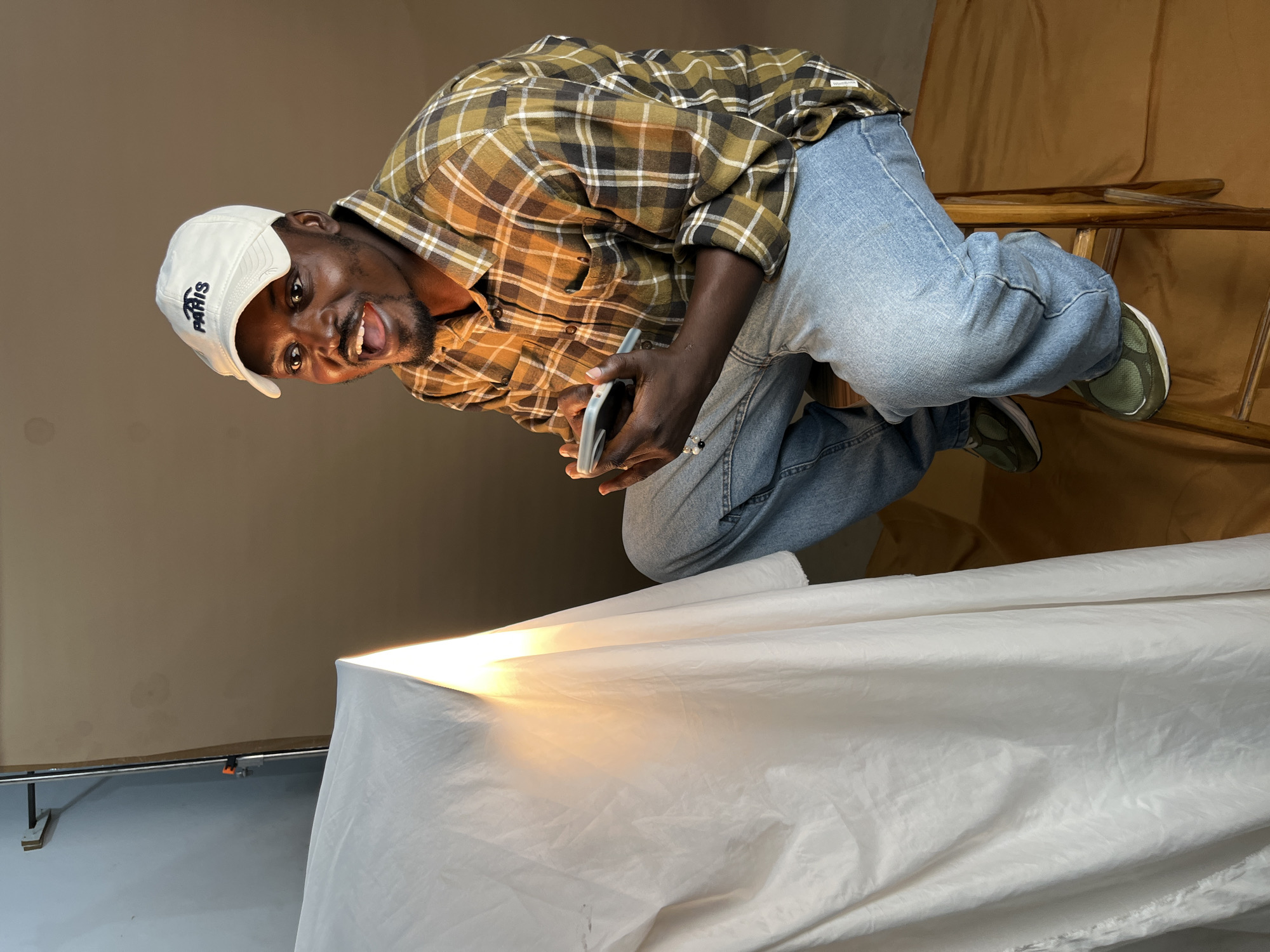 A young Black man wearing a white baseball cap with "PARIS" embroidered on it, a plaid long-sleeved shirt, and blue jeans is seated on a wooden stool. He is holding a mobile phone in his hands and looking off to the side with his mouth open in surprise or excitement. The man is positioned next to a large white cloth draped over an unseen object, with a bright light illuminating it from behind. The background is a plain brown wall. The scene appears to be a studio setup, possibly for a photoshoot, and is located in Gura Top, Nigeria.