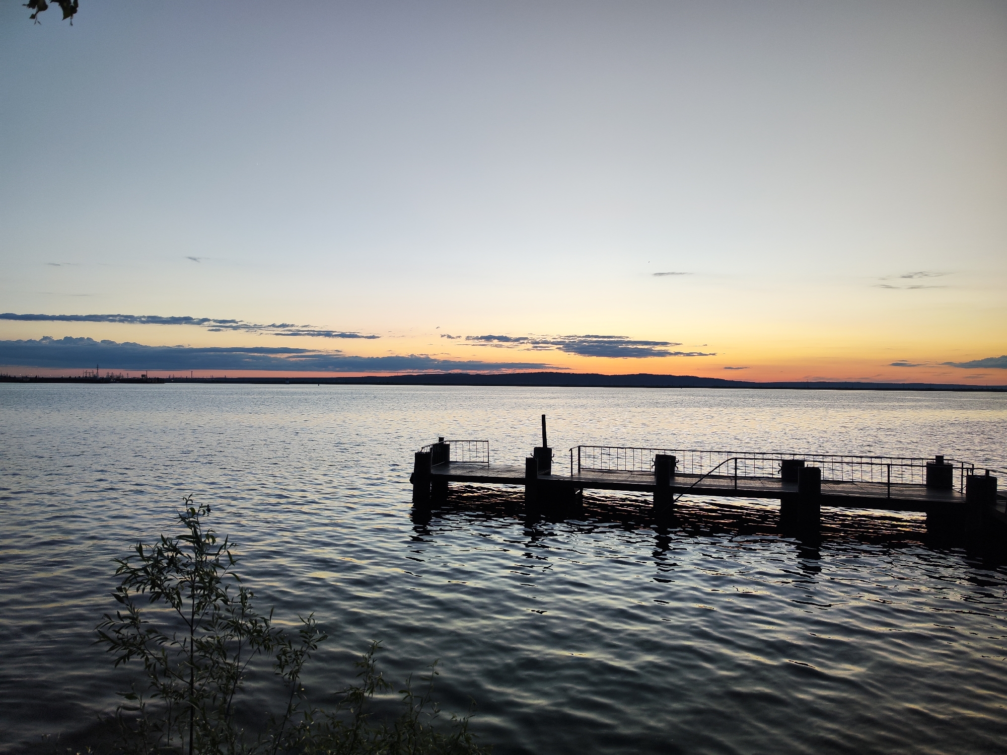 This wide-angle photograph captures a tranquil twilight scene over a large body of water, likely the Votkinsk Reservoir or Kama River near Chaikovsky, Russia. The sky, dominating the upper frame, transitions from a pale, cool blue overhead to soft oranges and yellows at the horizon, characteristic of either sunset or sunrise. Scattered horizontal clouds accentuate the horizon, reflecting the warm light.
A dark, narrow pier, constructed with what appears to be wood or concrete and featuring metal railings, extends from the right side of the frame into the gently rippling water. Its silhouette and those of its support posts are sharply defined against the water's surface, which subtly reflects the sky's colors. The water itself is calm, showing delicate wave patterns.
On the distant left shoreline, a dark silhouette of land reveals industrial structures, possibly factories or a power plant, contrasting with the natural landscape. The right distant shoreline presents a dark, tree-lined silhouette. In the immediate foreground on the bottom left, out-of-focus leafy vegetation, likely a bush or small tree, adds depth to the scene.
No specific activities or people are visible, contributing to the serene and contemplative atmosphere. The weather appears clear and calm. There is no visible text in the image. The overall impression is one of peaceful natural beauty at dusk or dawn, with a subtle indication of human development in the distant landscape.