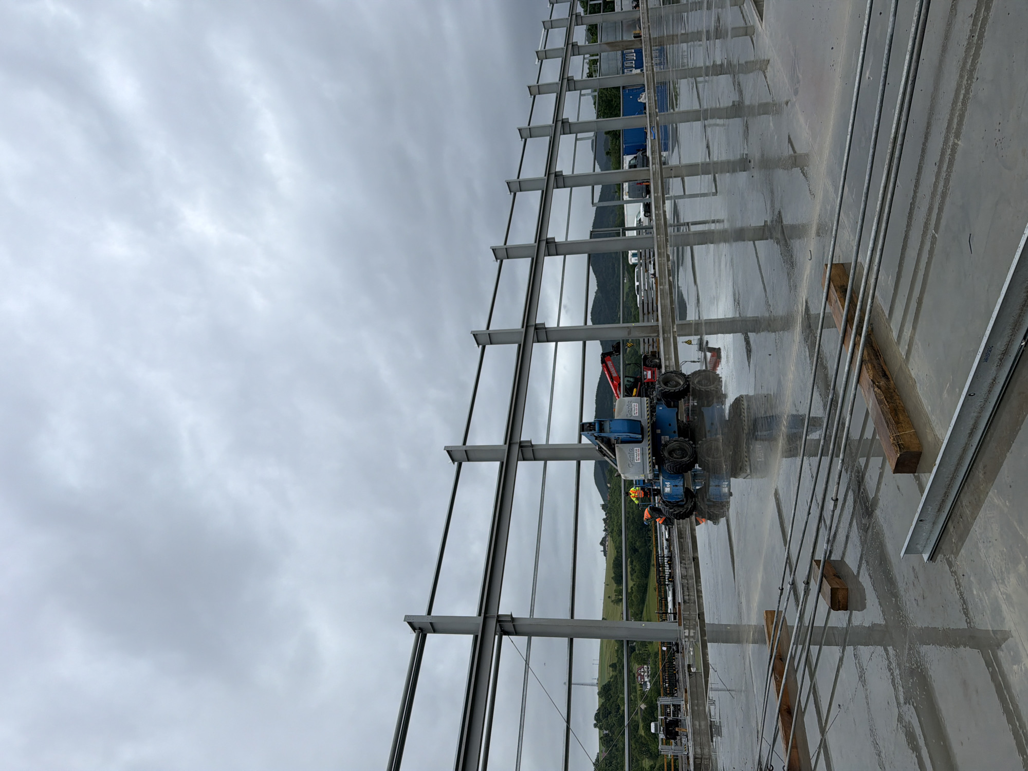 The image shows a construction site in Janowice, Poland, under a cloudy sky. The foreground features a wet concrete surface with various construction materials, including metal beams and wooden blocks, scattered across it. Reflections of the overhead steel structure and the wet surface are visible. In the mid-ground, a blue scissor lift with two construction workers in high-visibility vests is parked on the wet concrete. Further back, the skeletal structure of a large building, composed of steel beams and columns, is under construction. The background reveals a hilly landscape with green trees, some houses, and distant wind turbines under a heavily overcast sky. Several vehicles, including vans and a red telehandler, are also present on the site.