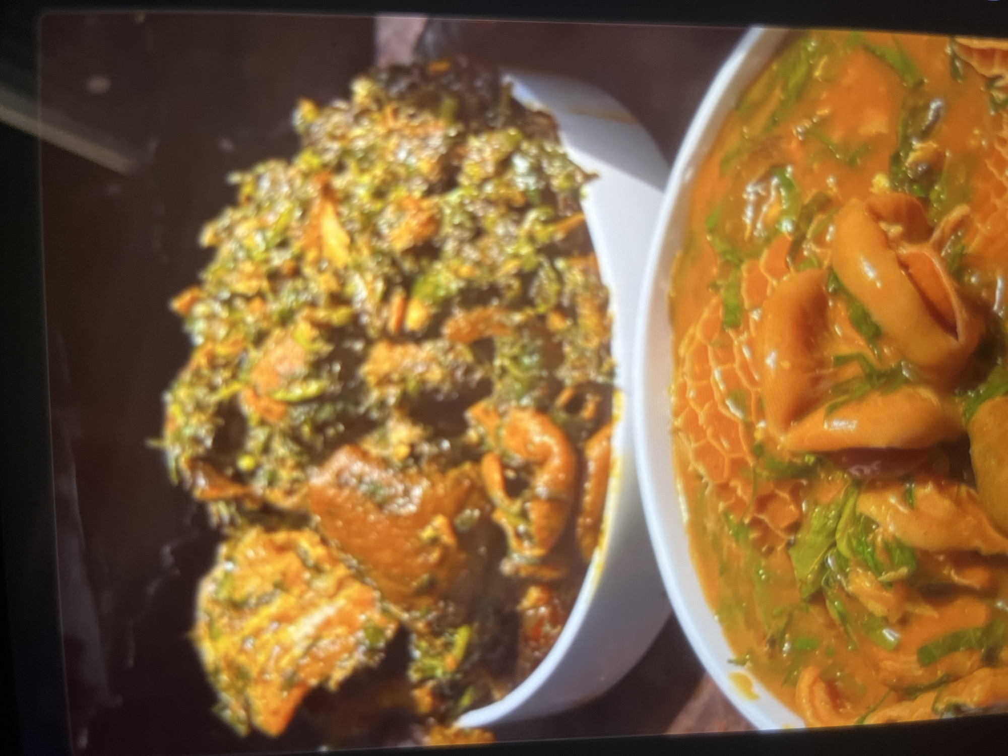 The image displays two white bowls of Nigerian cuisine. The top bowl contains a dish with leafy green vegetables, possibly efo riro, mixed with meat and a reddish-orange sauce. The bottom bowl features a hearty stew with large pieces of meat, likely goat or beef, in a thick, vibrant orange sauce, garnished with chopped green vegetables. The setting appears to be indoors, likely a restaurant or home in Abuja, Nigeria, given the presentation of the food. The lighting suggests a well-lit environment, focusing attention on the food. There are no people visible in the image.