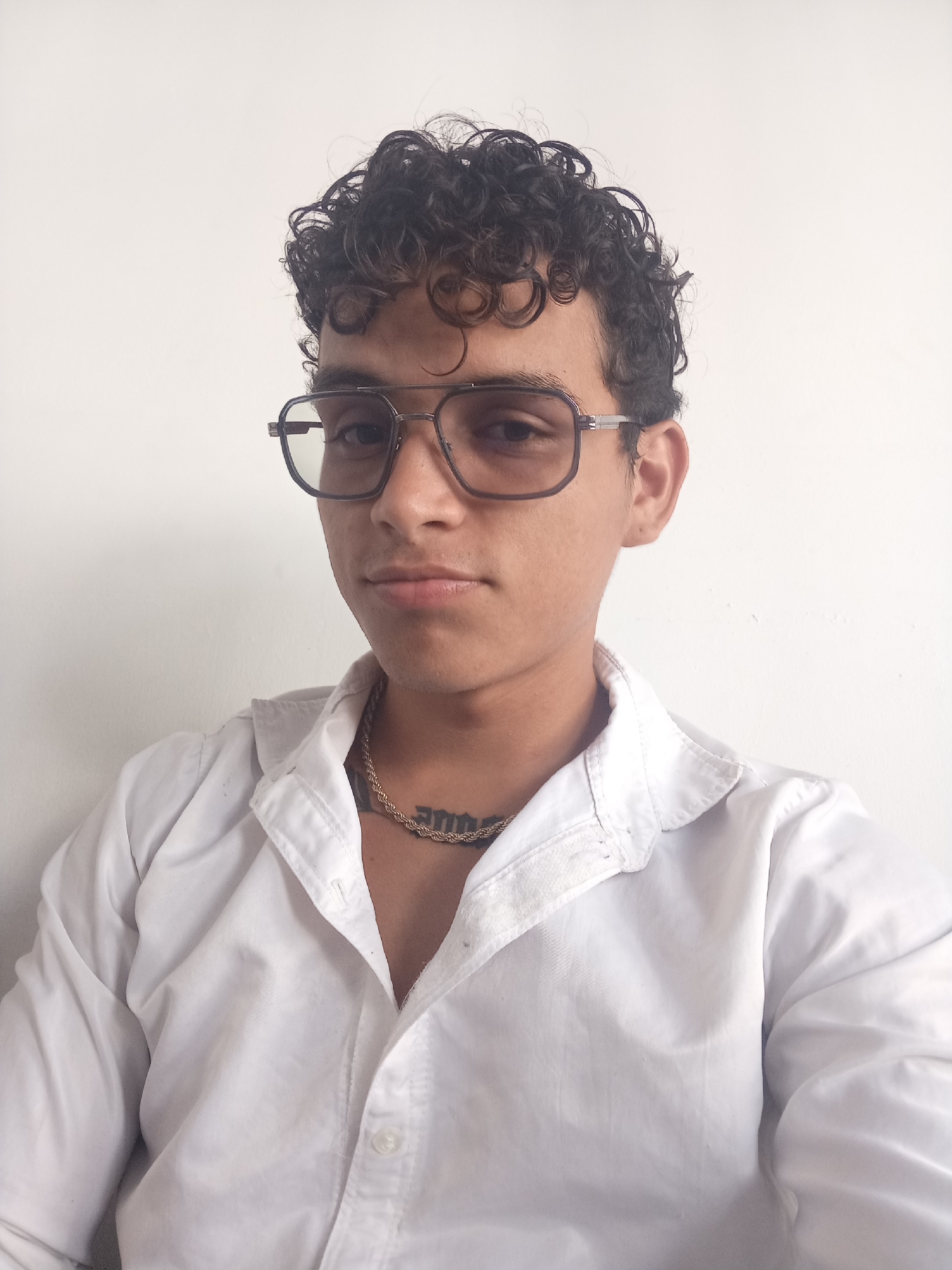 A young man with curly dark hair and glasses is shown from the chest up, wearing a white button-down shirt and a thick gold chain around his neck. A tattoo is visible on his chest, just above the chain. He is looking directly at the camera. The background is a plain white wall. The lighting is even and appears to be natural, casting soft shadows. This photograph was taken in Juan Griego, Venezuela.