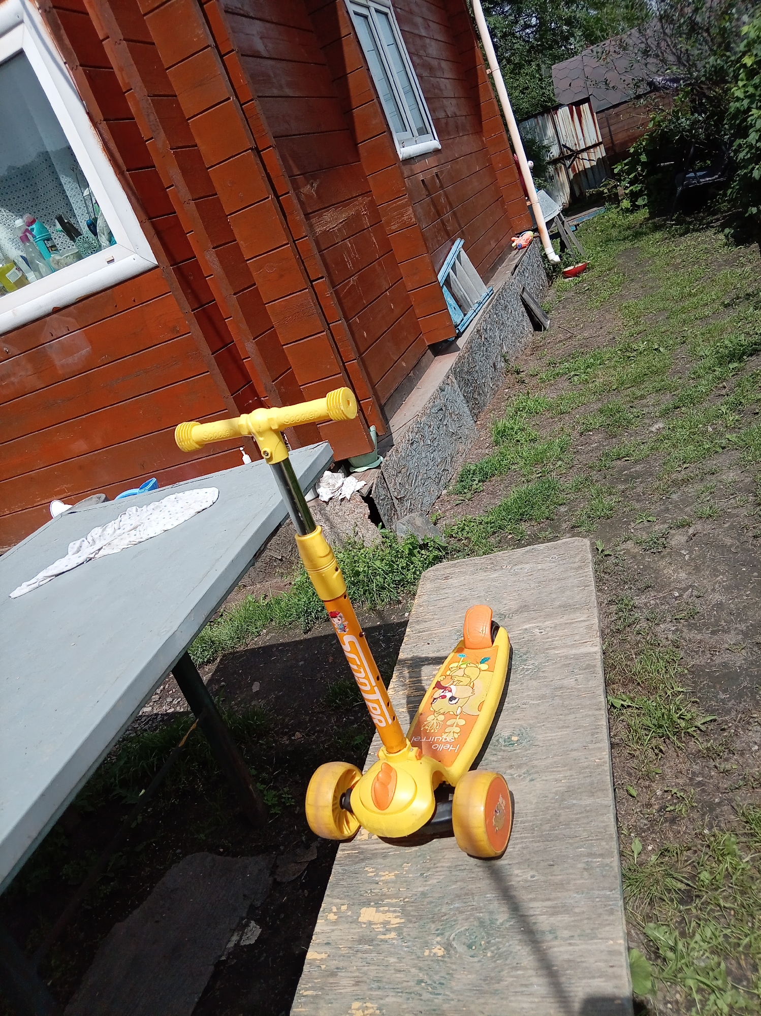 The image shows a yellow children's scooter on a wooden bench outdoors. The scooter has three wheels and a handle with grip. The deck of the scooter features graphics of cartoon characters, including squirrels, and text that appears to say "Hello Squirrel". In the background, a wooden building with a window is visible, along with a grassy area and some trees. The scene appears to be in a backyard or a similar outdoor setting. The lighting suggests it is daytime, with sunlight casting shadows. The location is identified as Artyom, Russia.