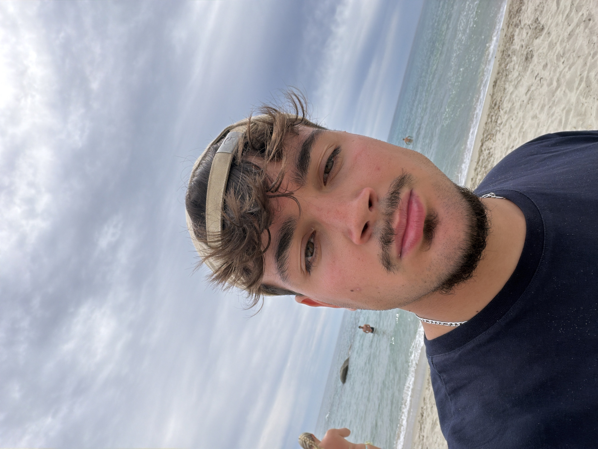 A man wearing a dark blue t-shirt and a light-colored baseball cap takes a selfie on a sandy beach. He has brown hair, a mustache, and a goatee. The ocean is visible behind him, with waves gently breaking on the shore. In the distance, two other people are in the water, one near a rock formation. The sky is overcast with clouds. The scene is consistent with a beach location in Tortolì, Italy.