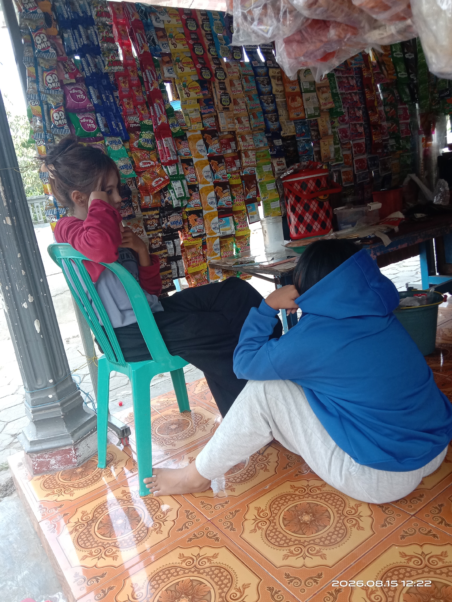 This image depicts an outdoor stall in Japanan, Indonesia, lined with an extensive display of packaged snacks and food items. Two individuals are present. On the left, a young person in a pink long-sleeved shirt is seated on a teal plastic chair, leaning on it with their arms and appearing to be resting their head on their hand. To the right, another individual is sitting on the tiled floor, wearing a blue hooded sweatshirt and light-colored pants. They are facing away from the camera, with their hands inside the hood of their sweatshirt. The stall itself is densely packed with rows of colorful snack bags, indicating a commercial setting. The ground is covered in ornate orange and brown tiles, and a grey pillar is visible on the left side of the frame.