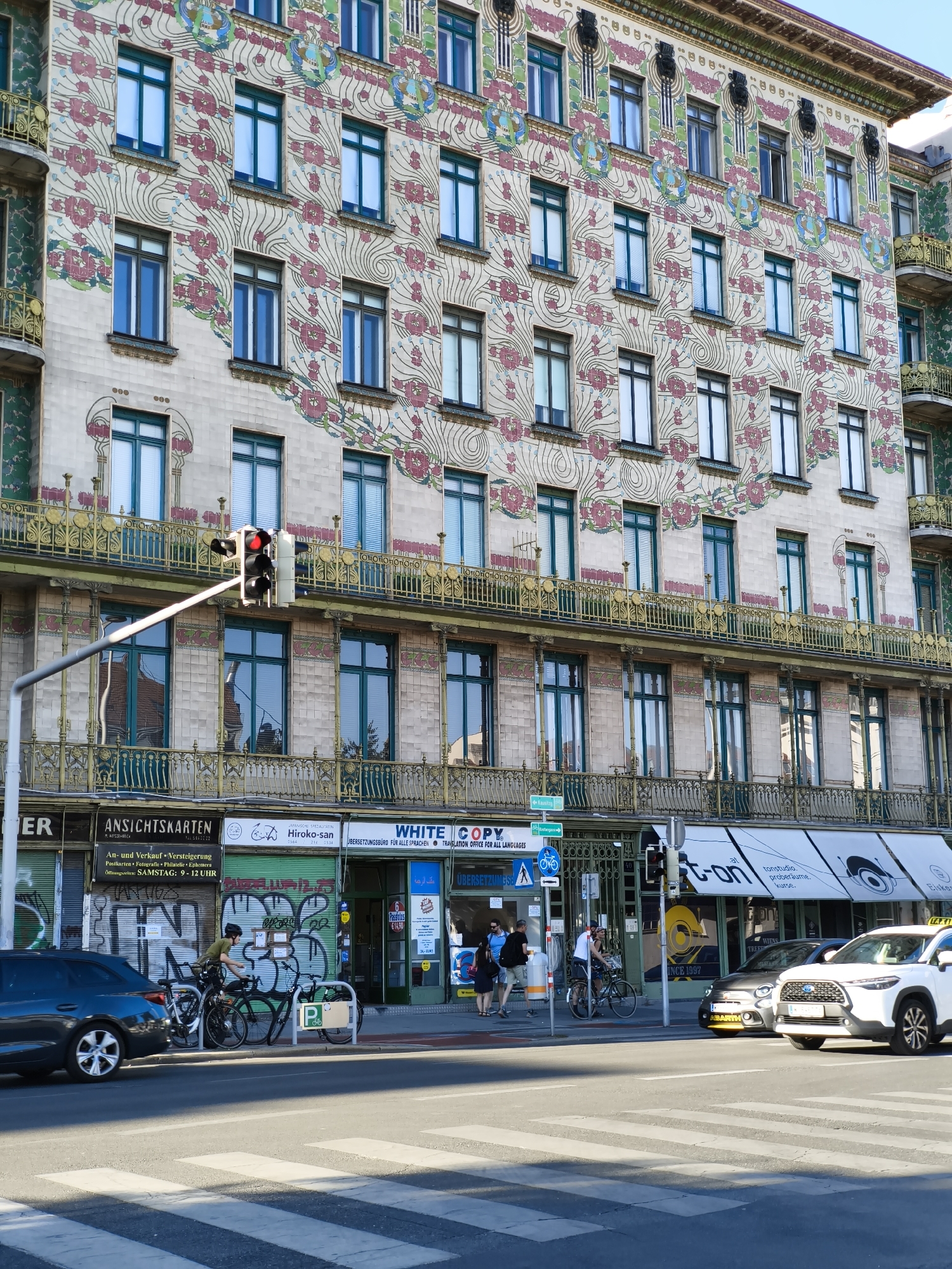 The image depicts a sunny street scene in Vienna, Austria, featuring the ornate facade of the Majolikahaus building. The building's exterior is adorned with a vibrant floral mosaic in shades of pink, green, and blue. On the street level, several shops are visible, including "An sichtskarten" and "WHITE COPY." A pedestrian crossing is in the foreground, with a traffic light showing red. Several people are present: a person on a bicycle is parked, another person is riding a bicycle, and pedestrians are seen near the shops. Two cars are on the road; one is dark-colored and the other is a white SUV. The sky is clear and blue.