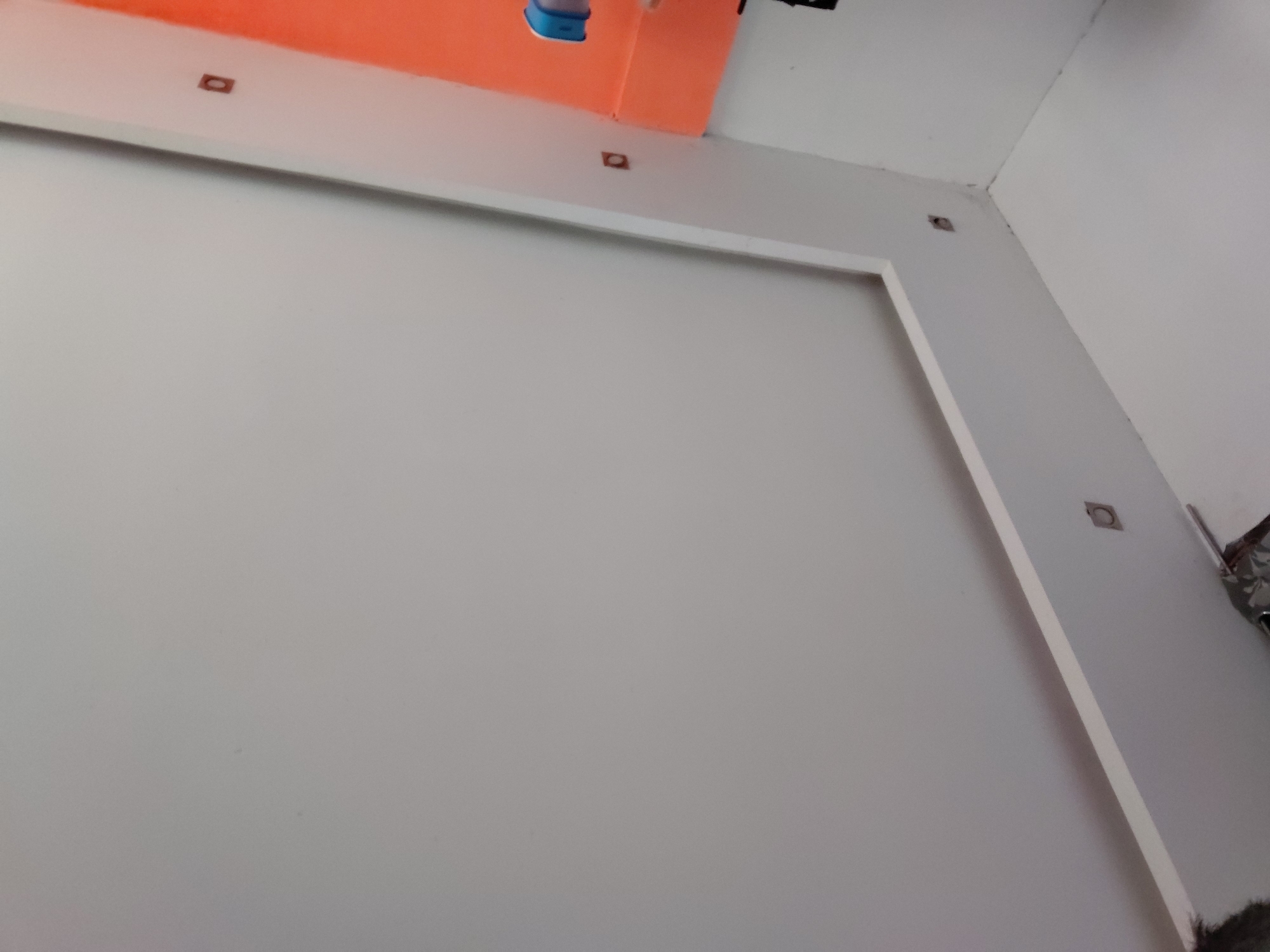 The image captures an interior scene, focusing on a wall arrangement within what appears to be a modern or recently renovated room. The dominant feature is a large, light gray wall, bordered by a prominent white rectangular frame that creates a recessed panel effect. Above this main wall, a section of the ceiling or an upper wall is painted a bright, vibrant orange.
Several small, dark, rectangular or square-shaped openings are visible. These are embedded into the white trim and on the adjacent white walls, appearing to be attachment points, electrical outlets, or conduits. One such opening is clearly visible on the white border against the orange section, and another on the white wall to the right. A blue object, possibly a plastic container or part of a fixture, rests on the orange surface in the upper left.
The room corners, particularly in the upper right, show clean lines where white walls meet the white ceiling. Towards the right side, there's a glimpse of a darker, textured object or area. The lighting is uniform, suggesting either bright artificial illumination or diffused natural light. No people, activity, or visible text are present in the image. The scene conveys a sense of a clean, minimalist design or an ongoing construction/renovation project in Santa Ana, Venezuela.