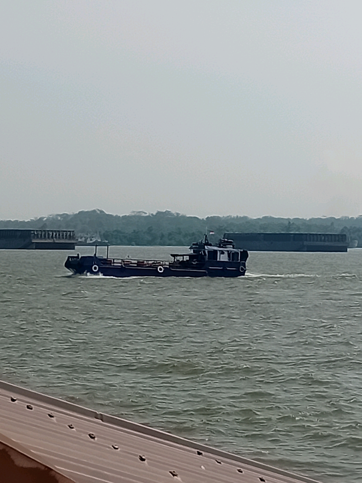 In Sungai Meriam, Indonesia, a navy blue cargo barge navigates on a body of water. The barge, with a dark gray railing and a flat deck, is moving towards the left of the frame, leaving a wake behind it. On the barge, there is a small cabin structure with a canopy and a flagpole. In the background, a long, dark, rectangular barge is stationary, and behind it, a lush green treeline stretches across the horizon under a hazy, light gray sky. In the foreground, the edge of a brown metallic surface with a patterned texture is visible, angled from the bottom left corner of the image.
