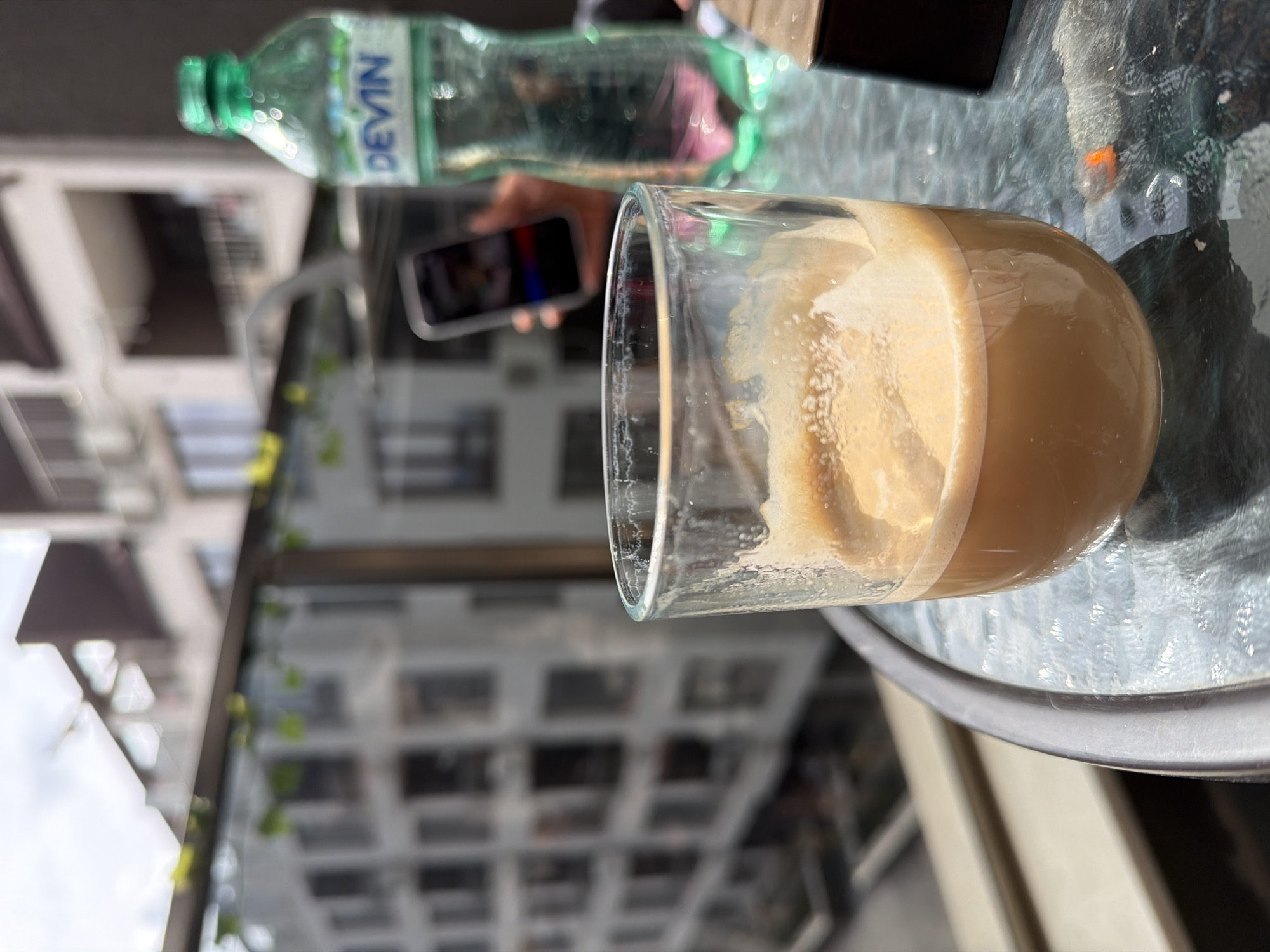 The scene depicts a clear glass containing a partially consumed, light brown beverage with foam, likely coffee, positioned on a textured glass-top outdoor table with a grey metallic rim. To the right of the glass, a clear plastic bottle of "DEVIN" brand mineral water, featuring a green cap and partially filled with liquid, stands upright. A hand holding a dark-colored smartphone, displaying a colorful image or interface, is visible behind the glass and bottle. A small, dark, rectangular object and an orange-tipped cigarette butt are also present on the table surface. The background is blurred, revealing modern, multi-story residential buildings with numerous windows and balconies, suggesting an urban environment. A glass balcony railing with small green leaves is visible in the mid-distance. The sky above is overcast. Based on the presence of the "DEVIN" brand water, the location is identified as an urban setting in Sofia, Bulgaria.
