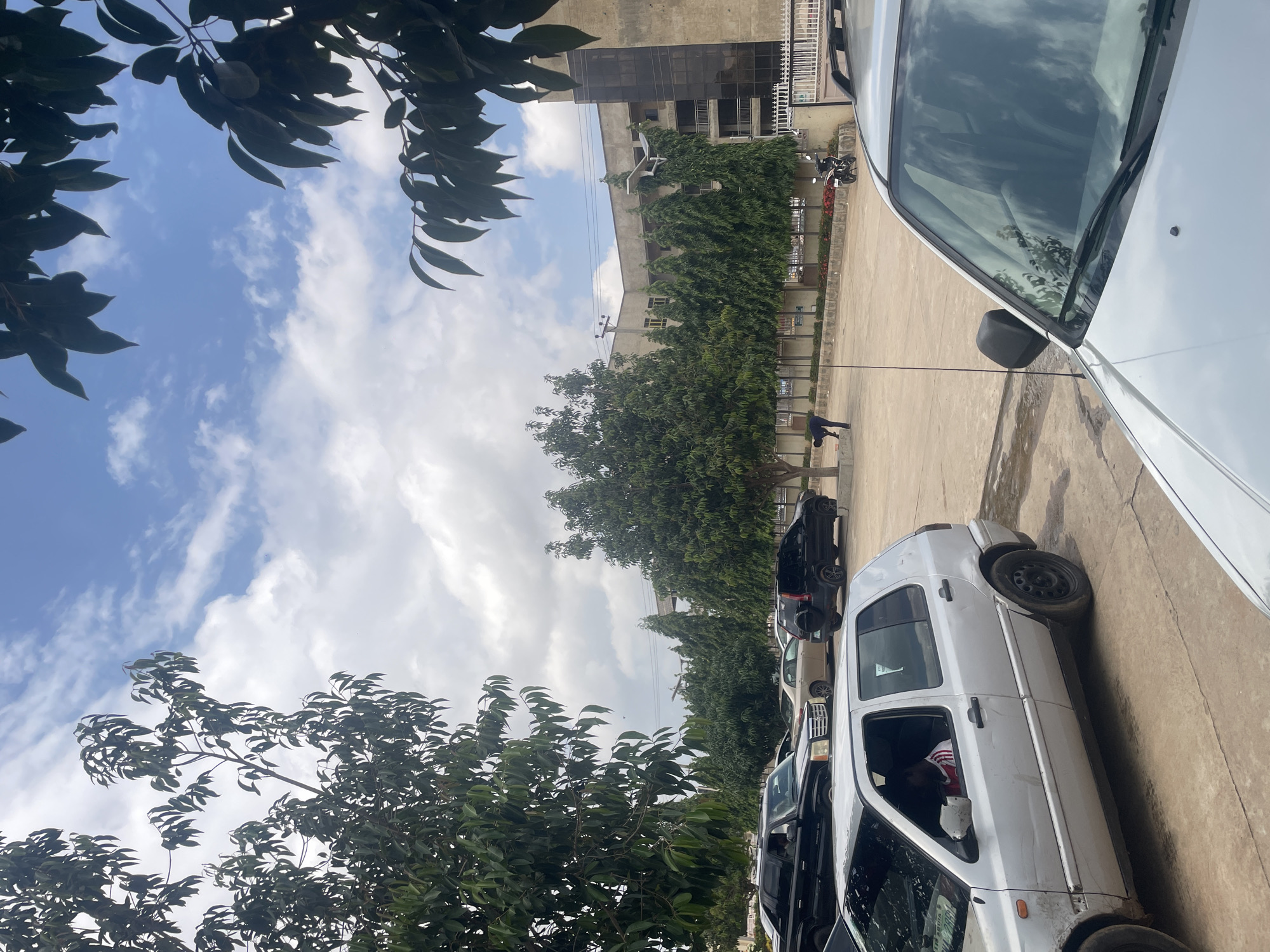 An eye-level, exterior, medium shot captures a paved outdoor area in Abuja, Nigeria, under a cloudy sky. In the foreground, the hood and windshield of a silver vehicle dominate the lower portion of the frame. To the left and slightly behind it, a silver Volkswagen Golf is parked, its driver visible inside, wearing a red and white striped shirt. A black SUV and a lighter-colored sedan are parked further back, with another black SUV to their right. In the middle ground, a person in dark clothing bends down on the pavement near a line of evergreen trees. Beyond the trees, a multi-story building with a beige facade and dark-tinted windows stands. A white metal fence lines the front of the building. A motorcycle is parked near the fence on the right side of the frame. Trees with dense green foliage frame the top and left sides of the image, with branches and leaves extending into the frame. The ground is concrete with some wet patches.