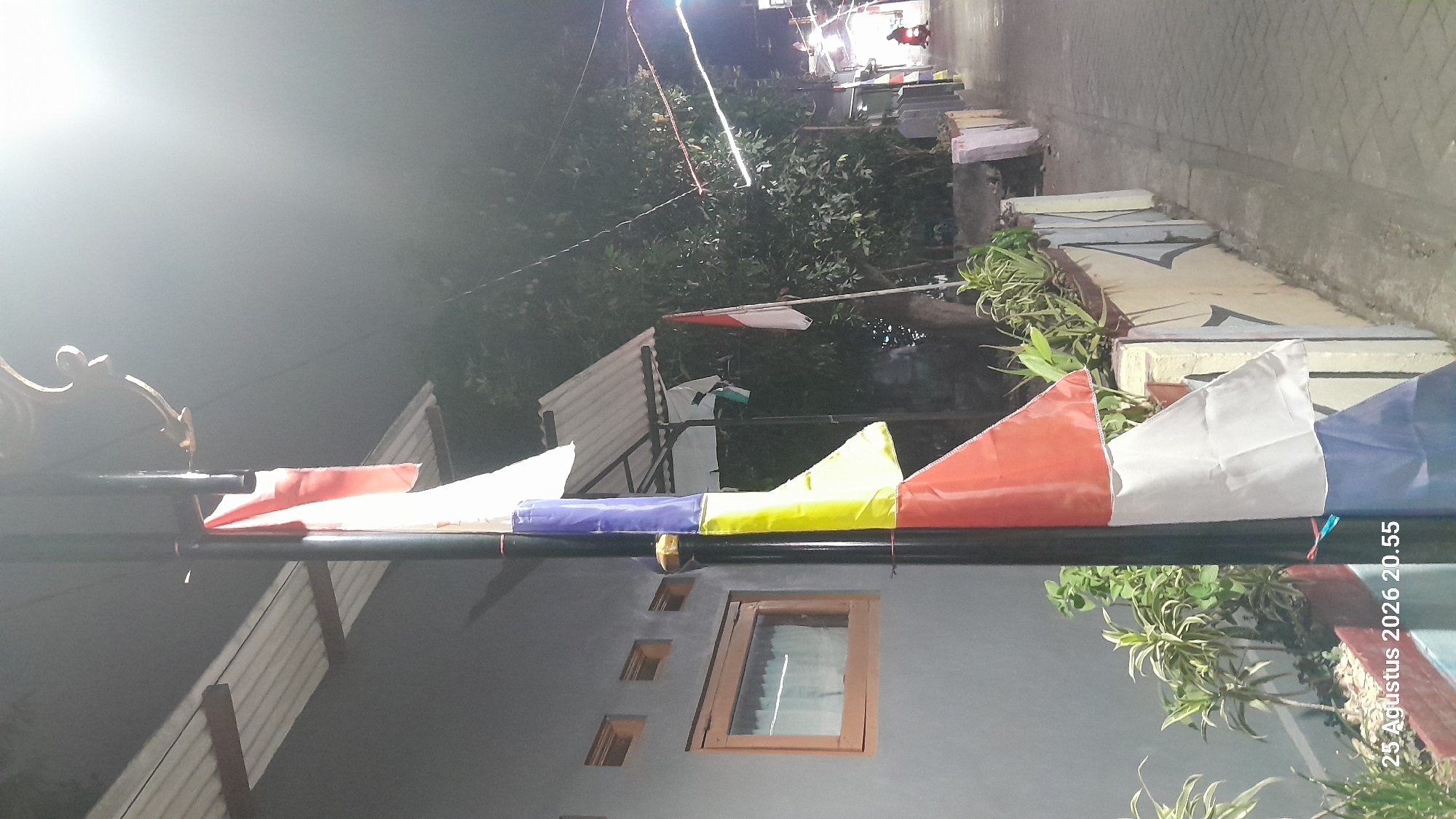 A vertical, low-angle, night shot captures a street scene in Madiun, Indonesia, illuminated by a bright light source above and to the right. The foreground is dominated by a black lamppost adorned with several Indonesian flags, red and white, and also blue, yellow, and white flags in a staggered arrangement. The flags hang limply, some partially obscured. To the left of the lamppost, a grey building with a corrugated metal roof is visible, featuring a window with a wooden frame. Along the right side of the frame, a paved walkway runs alongside a low decorative wall with geometric patterns. Beyond the wall, lush green foliage forms a dense backdrop. In the distance, a person on a red motorcycle is visible, moving along the street, and decorative lights are strung across the scene. The timestamp "25 Agustus 2026 20.55" is overlaid on the bottom left of the image.