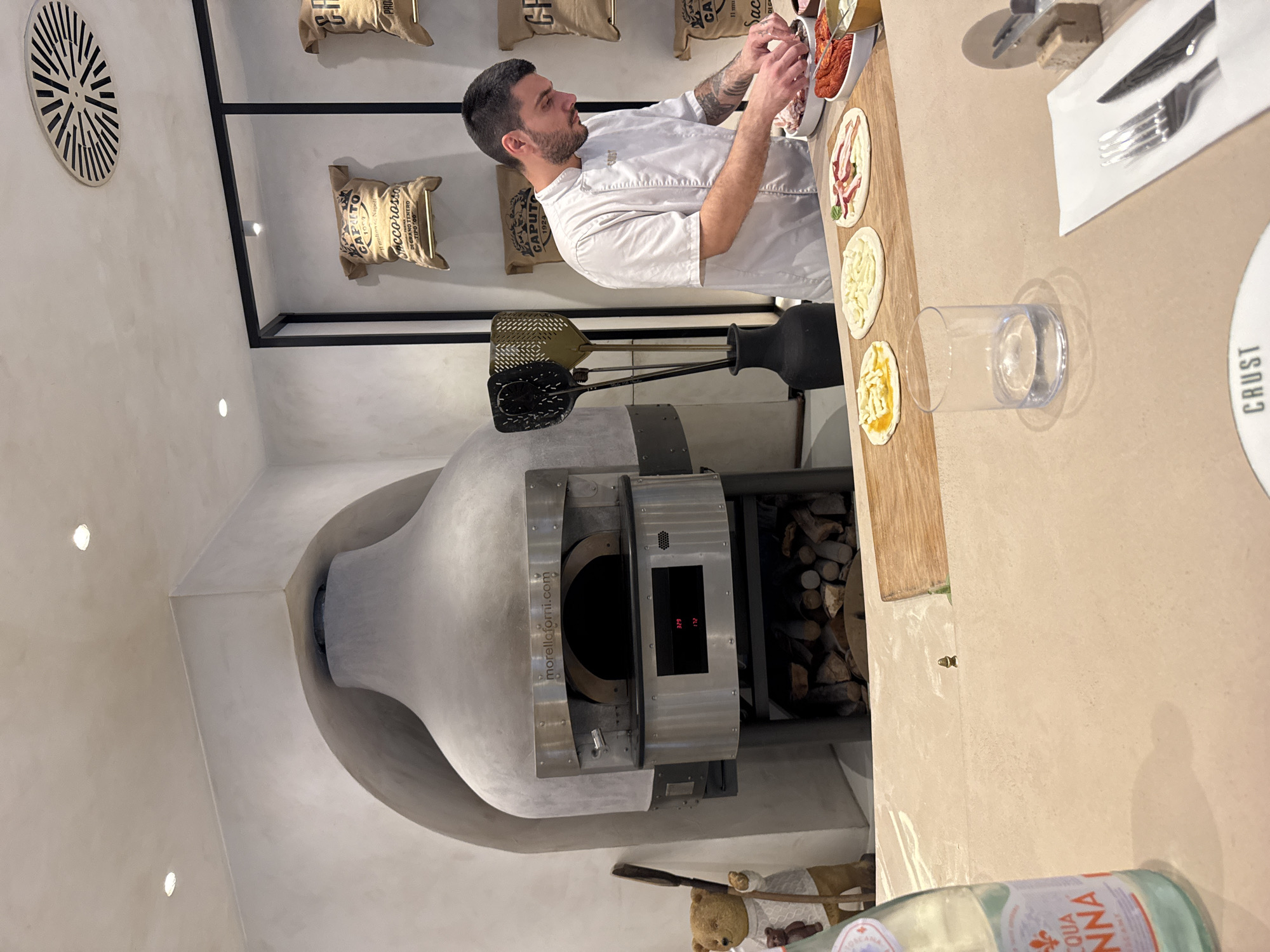 This media file captures a man in a chef's uniform meticulously preparing pizzas. He is in the process of adding toppings to small, individual pizzas, with various ingredients laid out in bowls in front of him. The scene appears to be inside a restaurant or kitchen, with a prominent wood-fired pizza oven dominating the background. The oven is a focal point, made of a light-colored material with metallic accents and a digital display showing temperature readings. Above the oven, sacks labeled "CAPUTO" and "accorosso" are displayed, suggesting they contain flour or other pizza-making ingredients, possibly hinting at the restaurant's commitment to quality sourcing.
Several pizza-making tools, including peelers, are neatly arranged near the oven. On a wooden countertop in the foreground, a few small pizzas are already assembled, awaiting their turn in the oven, alongside a clear glass and a bottle of Acqua Panna water. A close-up crop reveals the text "CRUST" on a white plate, which may be the name of the establishment or a specific menu item. The overall impression is one of a professional and artisanal approach to pizza making. The lighting is somewhat dim, suggesting it could be evening or a dimly lit interior. The presence of the pizza oven and the focus on pizza preparation strongly indicate a culinary setting.