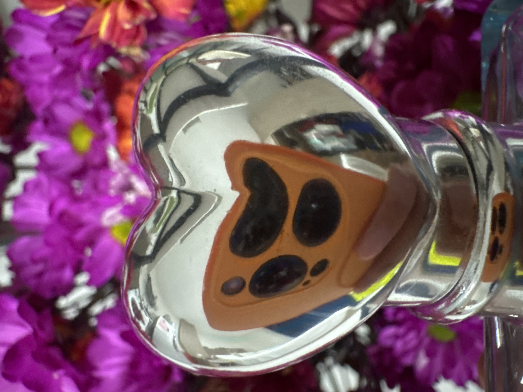 The image shows a close-up of a silver heart-shaped object with a highly reflective surface. The surface reflects an orange object with several black circular elements, likely a mobile phone with multiple cameras. The heart-shaped object is positioned in front of a blurred background of bright purple and pink flowers, with some yellow and red accents. The bottom of the heart-shaped object appears to be attached to a metallic stand or base. There is no identifiable location in the city of Nha Trang Ward, Vietnam within the image. The scene is static and focuses on the decorative object and its reflection.