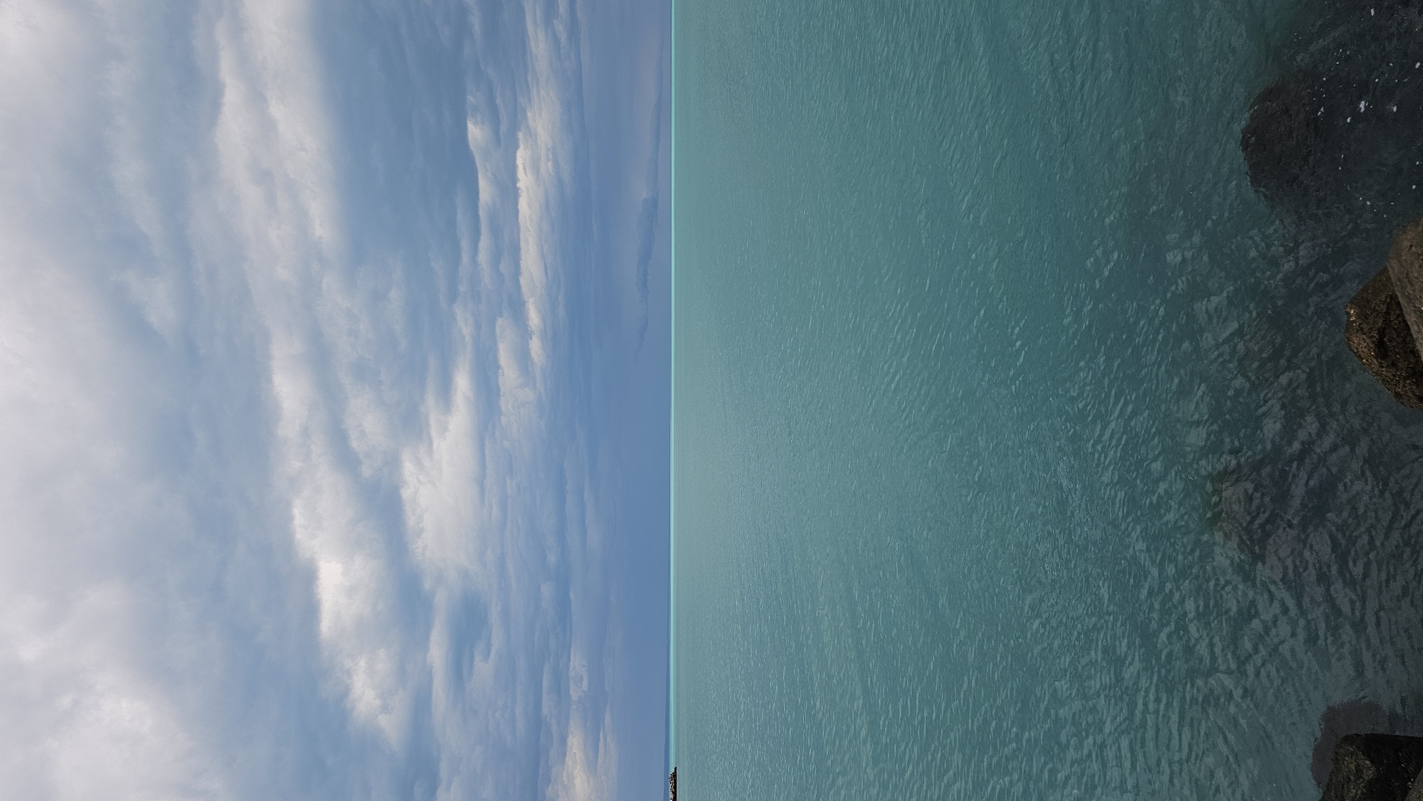 The image depicts a serene coastal scene with a clear division between the sky and the sea, likely taken at or near a beach. The left side showcases a bright blue sky filled with streaky white clouds. On the right side, the turquoise-colored water is visible with a gentle ripple effect on its surface. Along the right edge of the image, there is a dark rocky area, most likely a part of a breakwater or a rocky coastline.
While the image is a common landscape of sea and sky, the presence of the rocky coastline suggests that the photograph was probably taken at a beach.
However, without more context, the exact location within Varna cannot be precisely pinpointed.