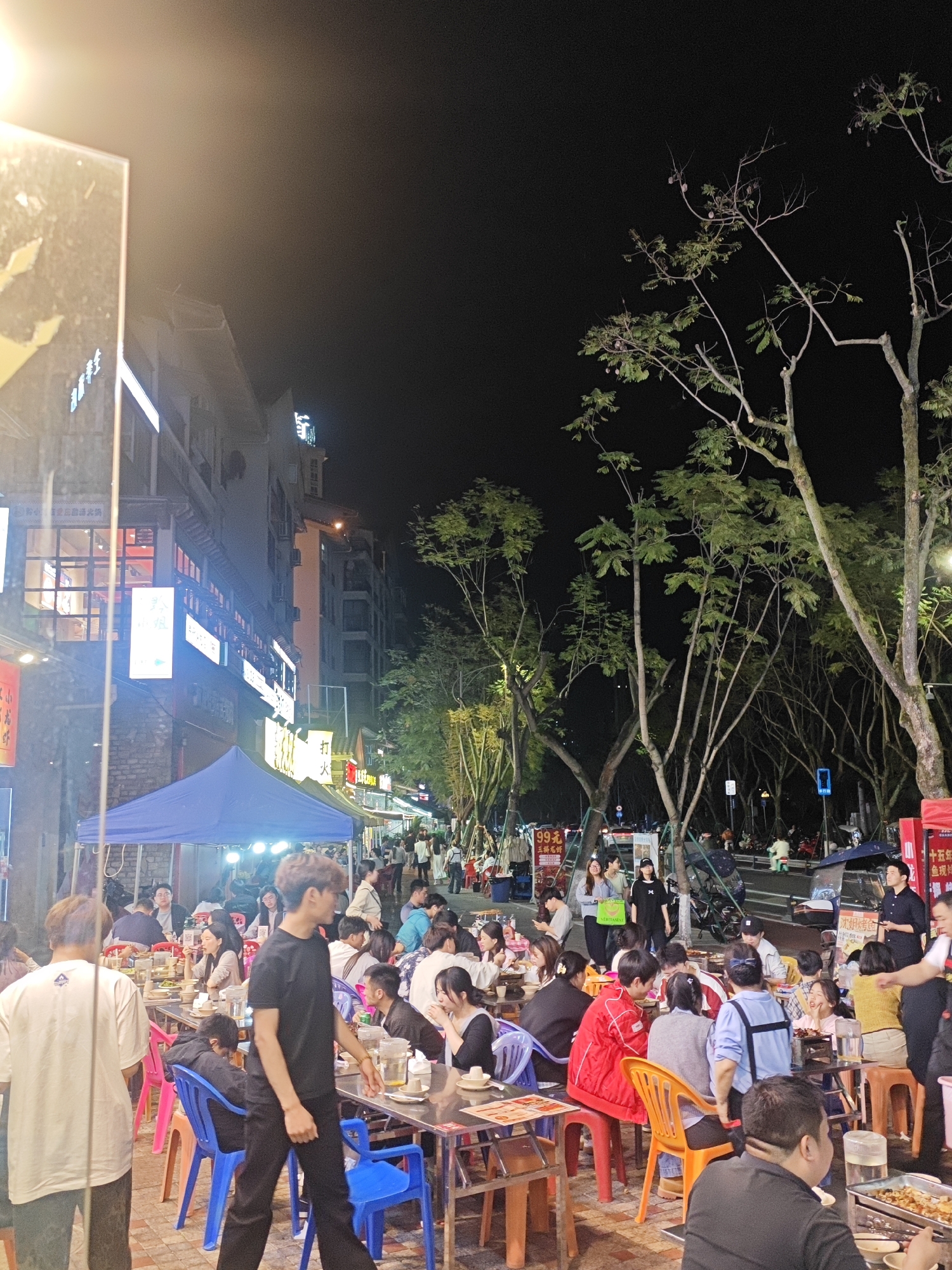 The image depicts an outdoor street food scene at night in Chongqing High-tech Zone, China. Multiple tables and chairs are arranged along the sidewalk, occupied by numerous people dining. The people are mostly dressed in casual attire. Street vendors and waiters are serving and attending to the customers. A blue tent is set up near the building on the left side of the street, and several trees line the street on the right. Streetlights illuminate the area, and buildings with illuminated signs are visible in the background. The overall atmosphere is lively and bustling.
