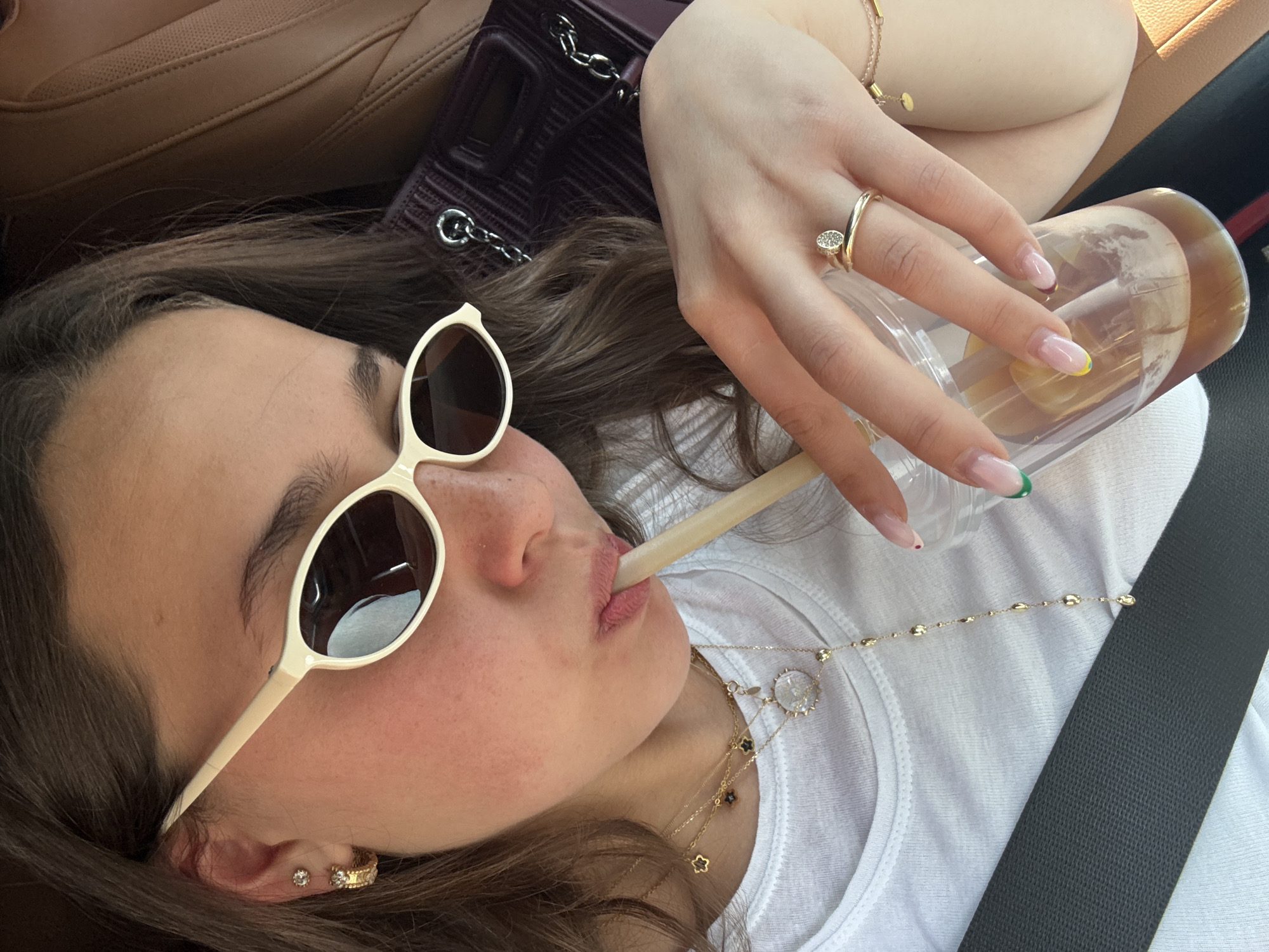 The image depicts a close-up, high-angle shot of a young woman inside a car, with the location context provided as Yerevan, Armenia. She is seen from a diagonal perspective, showcasing her upper body, head, and left arm.
The woman has long, dark brown hair and is wearing cream-colored, oval-shaped sunglasses. She is in the act of drinking from a clear plastic cup filled with an amber-colored liquid, using a light-toned straw. Her left hand holds the cup, revealing a gold ring on her ring finger with a small, sparkly embellishment, possibly resembling a nail-head design. Her fingernails have a French manicure style with a clear base, white tips, and colorful accents (green, yellow, and a small red/orange dot) on some tips. A delicate gold bracelet with small charms is visible on her left wrist.
She is dressed in a white ribbed t-shirt, over which several gold necklaces are layered. These include a chain with a prominent circular pendant featuring a star-like motif, another with small four-leaf clover-shaped charms, and a simpler chain with small beads. A dark gray seatbelt is fastened across her chest.
In the background, elements of the car's interior are visible: tan leather upholstery of a car seat and a dark-colored, possibly burgundy or deep purple, quilted handbag with a silver chain strap. The bright lighting suggests it is daytime. The overall impression is one of a casual, relaxed moment, possibly while traveling as a passenger. No visible text is present in the image.
