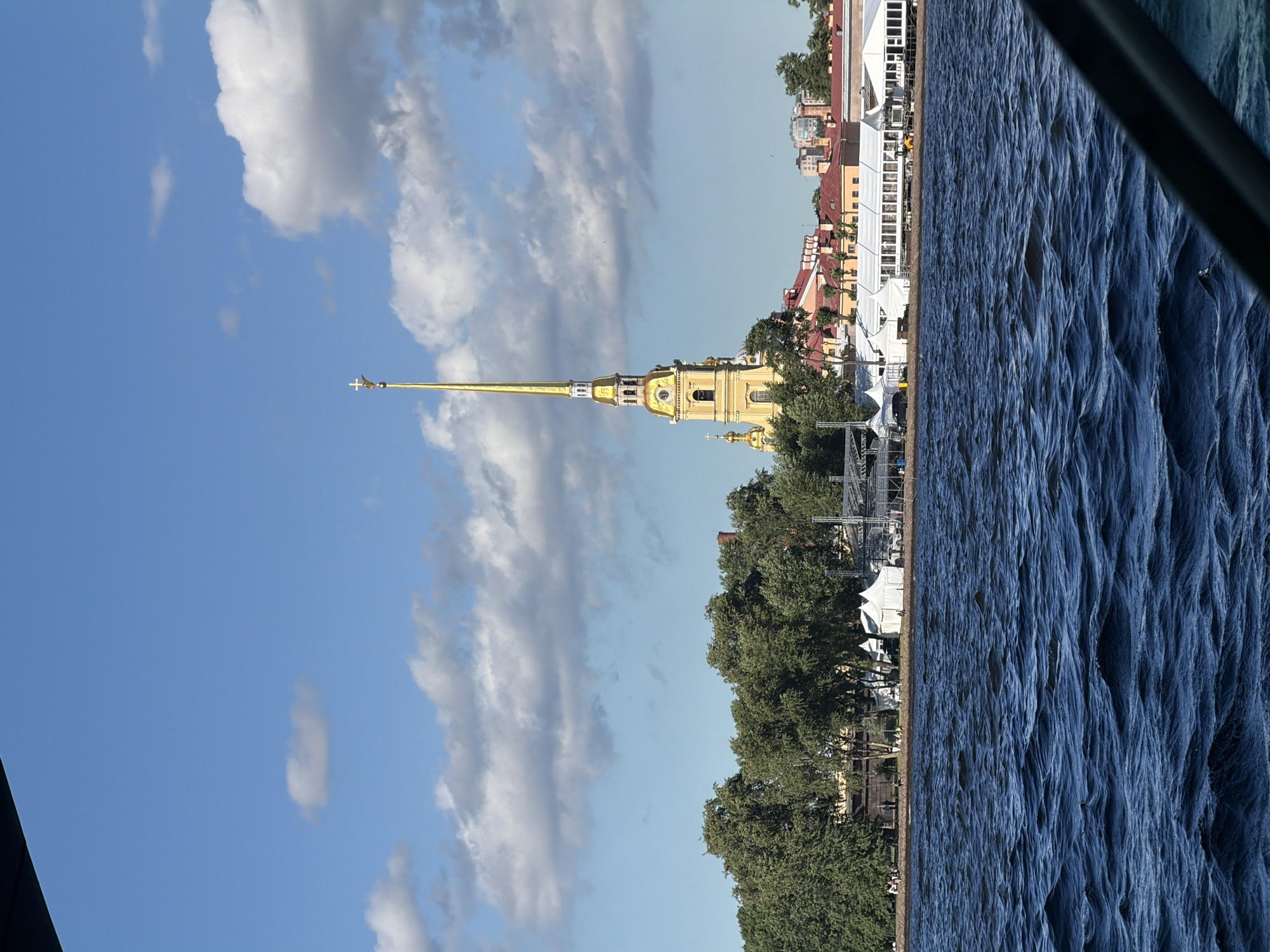 A body of water with choppy waves is in the foreground. In the mid-ground, a shoreline with lush green trees is visible, behind which are several white tents and some buildings with red roofs. Dominating the mid-ground is a tall, slender yellow tower with a golden spire topped with a cross. The sky above is a clear blue with scattered white clouds.