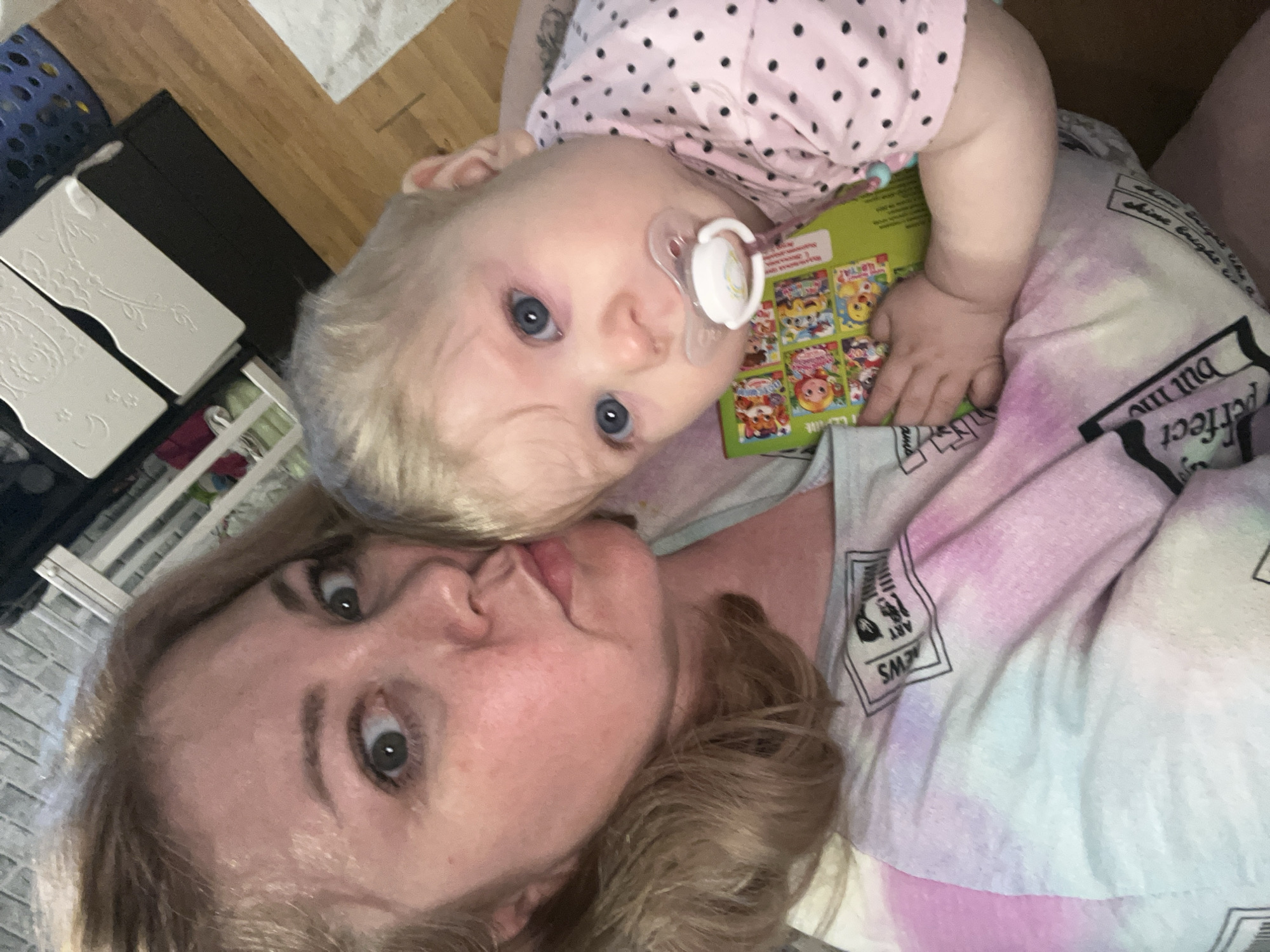 A woman and a baby are lying down, presumably indoors. The baby is wearing a pink polka dot shirt and has a pacifier in their mouth. The woman has blonde hair and is making a kissing face towards the camera. The baby is holding a colorful book. The woman is wearing a tie-dye shirt with text on it. A blue laundry basket and a white dresser are visible in the background. The location in the city of Vesyoloye, Russia, cannot be determined from the image.