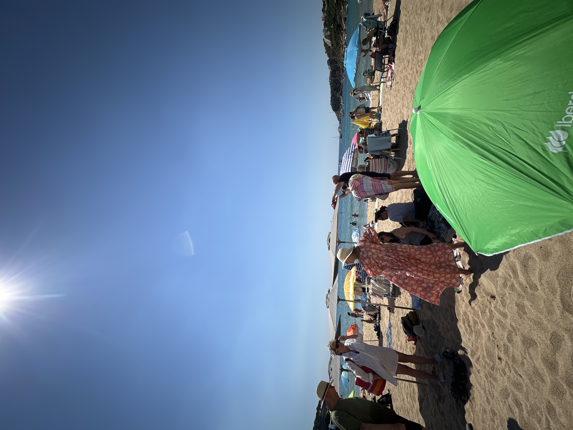 This is a medium-shot, eye-level, vertical, outdoor, daytime photograph taken at the beach in Sant Feliu de Guíxols, Spain. The sun is bright and high in the sky, casting strong shadows on the sand. The beach is populated with people enjoying the sunny weather. In the foreground, a large green umbrella is prominent, its logo partially visible. To the left of the umbrella, several people are standing and sitting on the sand, some with umbrellas providing shade.
Further back, more beachgoers are visible, some relaxing under umbrellas, others in the shallow water. The water is calm and blue, with a few boats anchored offshore. The background shows a hilly coastline with buildings and vegetation. The scene captures a typical summer day at the beach, with people engaged in various leisure activities.