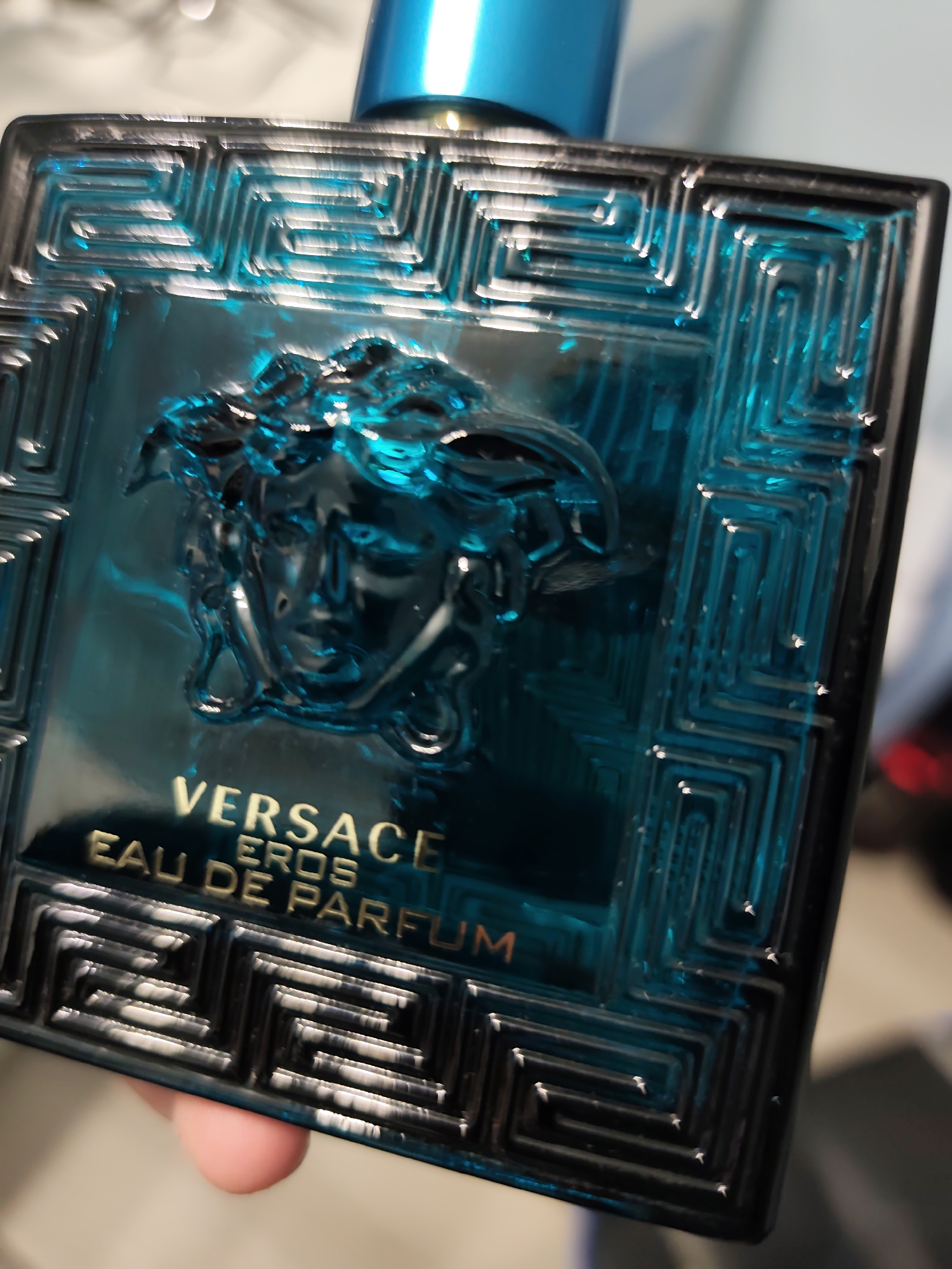 A close-up, slightly angled photograph depicts a bottle of Versace Eros Eau de Parfum. The bottle is rectangular, made of deep teal or turquoise glass, and features a dark, likely black, frame around its edges. A prominent raised Medusa head logo, crafted from the same teal glass, is centered on the front surface. Surrounding the Medusa head and bordering the bottle is an intricate, embossed Greek key (meander) pattern, which appears highly reflective, catching ambient light and highlighting its geometric lines. Below the Medusa head, gold-colored text is visible: "VERSACE" in a larger uppercase font, followed by "EROS" in a slightly smaller uppercase font, and then "EAU DE PARFUM" in a smaller uppercase font. The upper portion of a bright blue, cylindrical cap is visible at the very top of the bottle. The bottom edge of the bottle is partially held by a person's light-skinned finger, which is visible at the lower edge of the frame. The background is significantly blurred and out of focus, appearing as soft, light tones, indicating an indoor setting with ambient lighting that creates prominent reflections on the bottle's glass surfaces.