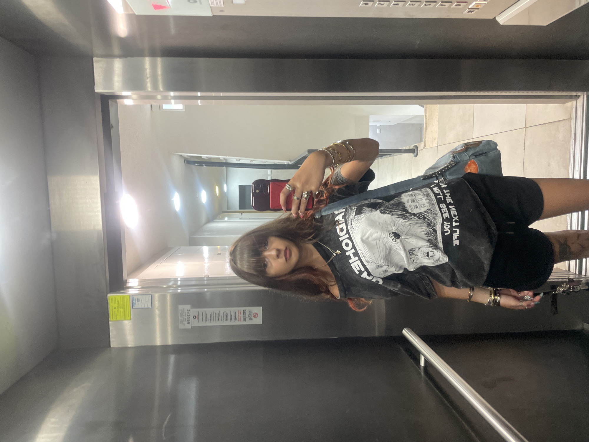 A young woman with long, dark hair and bangs is standing in an elevator, taking a mirror selfie with her red smartphone. She is wearing an off-the-shoulder graphic t-shirt and black shorts. She has several bracelets and rings on her left hand and a necklace with a pendant. A denim bag is slung over her left shoulder. The elevator has stainless steel walls and a metal handrail. A sign with Hebrew text is visible on the wall of the elevator. The floor of the elevator appears to be tiled. The background of the selfie shows a hallway with lights and doorways. The image appears to have been taken in Raanana, Israel, likely in a residential building or a commercial space.