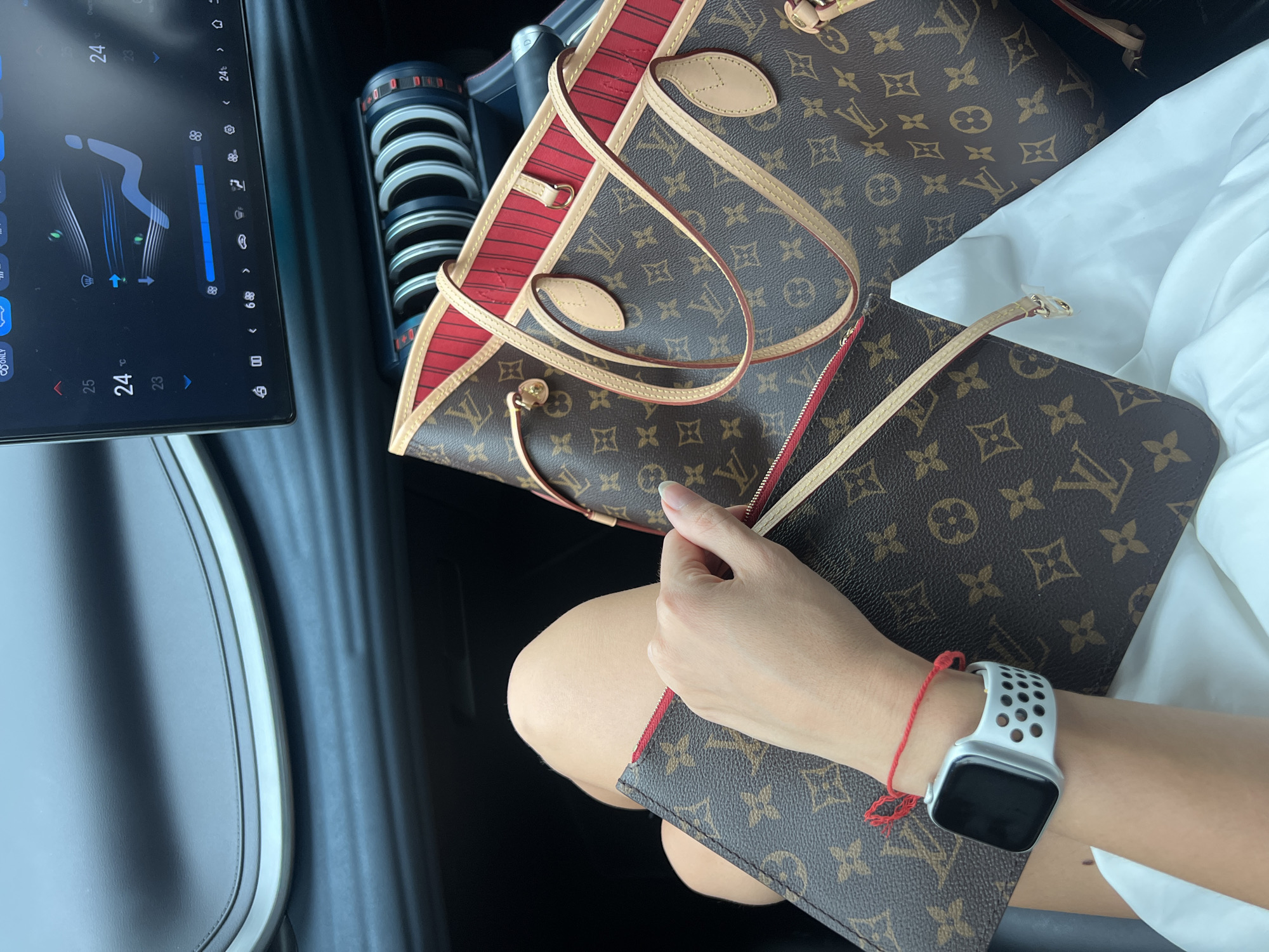 The image depicts the interior of a vehicle, likely a car, with a focus on the passenger side. A Louis Vuitton Neverfull bag with a red interior trim and a matching pouch rests on what appears to be a white blouse or fabric on the passenger's lap. The passenger is visible by their lower arm and hand, where an Apple Watch with a white sport band and a red string bracelet are worn. The central console of the car includes a large display screen showing climate control settings. There are also cylindrical objects in a row.