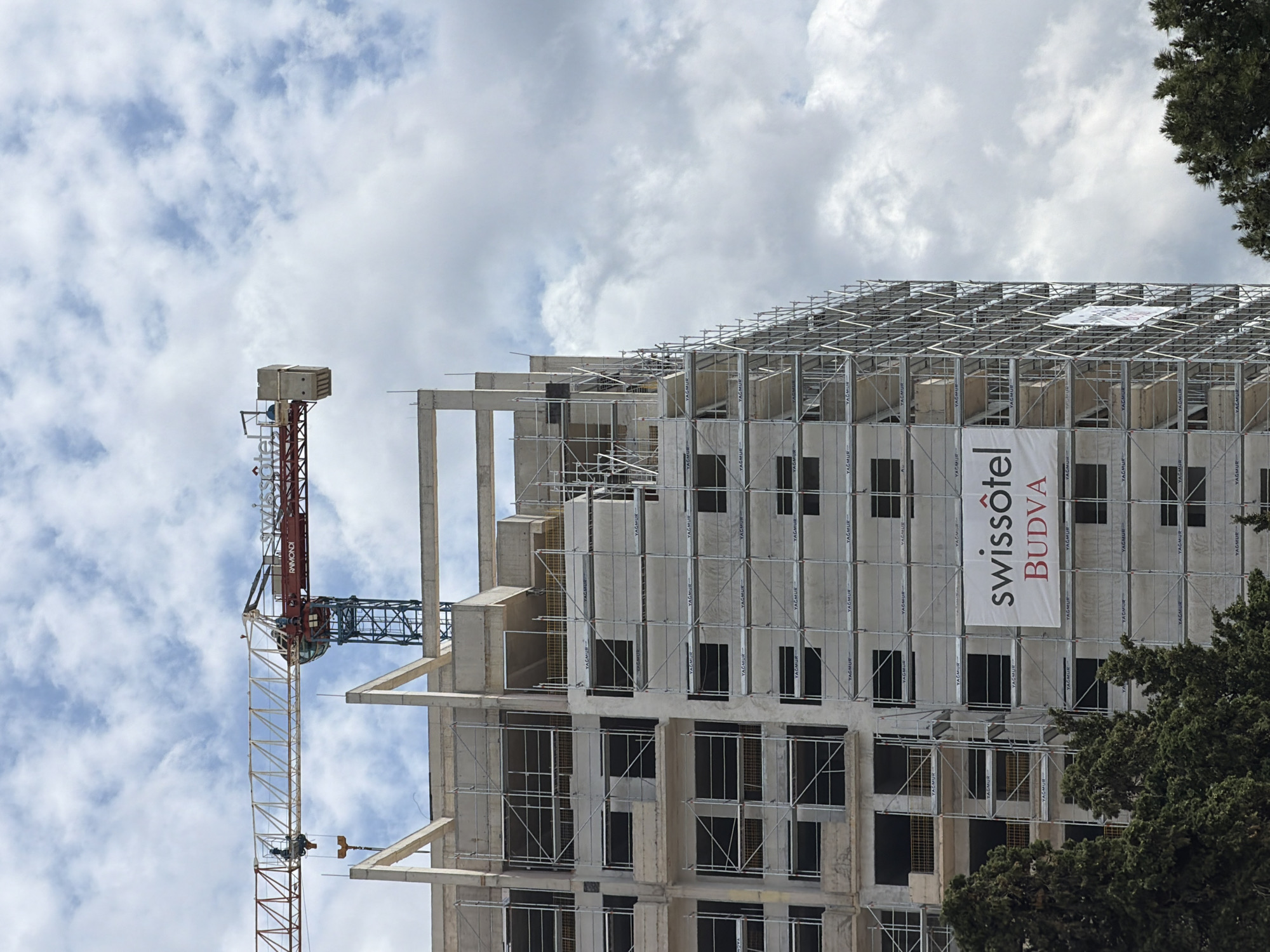This daytime photograph captures a large building under construction in Budva, Montenegro. The central focus is a multi-story concrete-framed structure, currently in an unfinished state with its skeletal frame of columns and beams clearly visible. Extensive scaffolding, with the word "YAĞMUR" repeated numerous times on its components, covers significant portions of the building's facade.
A large white banner, prominently featuring the text "swissôtel BUDVA" in black and red lettering, is attached to the building, indicating its future purpose as a hotel. To the left of the building, a tall tower crane with a red and white lattice structure is actively engaged in the construction process. The name "RAIMONDI" is visible on the crane's jib.
The sky above is filled with white and light grey clouds. On the far right side of the image, the dark green foliage of several trees is visible, partially framing the construction site. The overall scene depicts an active construction environment, marked by the presence of heavy machinery and scaffolding. No people or specific activities are discernible.