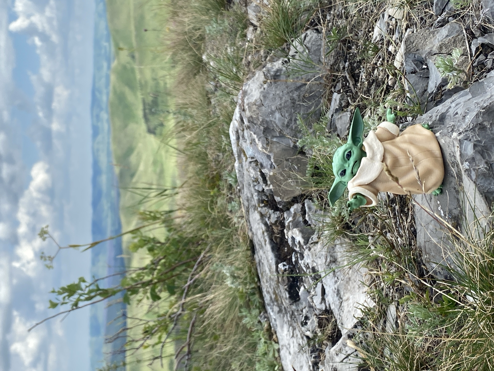 An outdoor, eye-level, medium shot captures a miniature toy of the character "The Child" (Grogu), also known as Baby Yoda, placed on a rough, gray, rocky surface. The toy is positioned in the lower right quadrant of the frame, facing slightly to the left. It is depicted wearing its characteristic tan robe. The terrain around the toy is uneven and covered with dry grass and small green plants. Behind the immediate foreground, a large, irregularly shaped gray rock occupies the center and left side of the image. In the background, rolling green hills stretch into the distance, under a partly cloudy sky with large, white cumulus clouds against a pale blue sky. The overall impression is of a scenic, natural landscape. The location of Абзаново, Russia, is not visually evident from the image itself, and can only be inferred from external information.