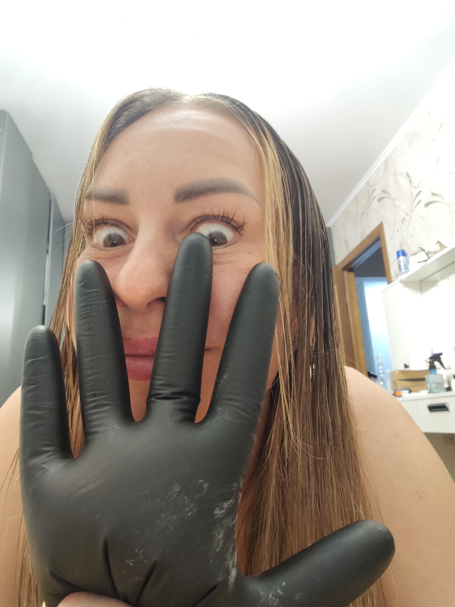 The image shows a close-up of a woman's face and hand, taken from a first-person perspective. She is wearing a black glove on her right hand, which is held up in front of her face, partially obscuring it. Her eyes are wide open and looking directly at the camera, with an expression that could be interpreted as surprise or playful. Her hair is long and appears to be wet or recently styled, with streaks of blonde mixed with a darker color.
The background suggests an indoor setting, likely a bathroom or a dressing area. To the left, a gray closet or cabinet is visible. To the right, a doorway leads to another room, and a white shelf with various items, including bottles and containers, is present. The wall in the background has a patterned wallpaper.
The image does not contain any discernible features that would place it in Almaty, Kazakhstan, or any other specific city. The setting appears to be a private residential or salon space.