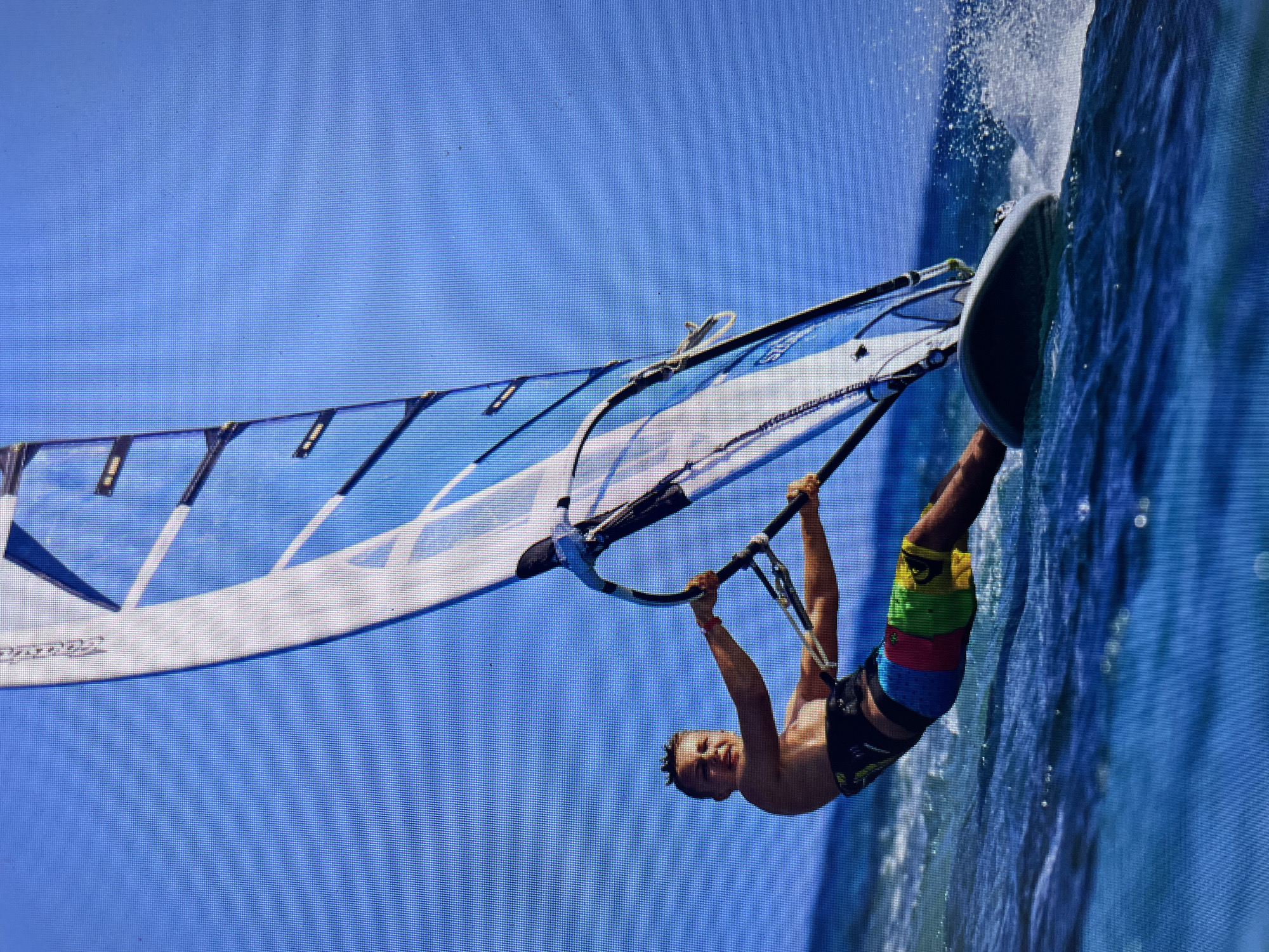 A man is windsurfing on blue water under a clear blue sky. He is gripping the boom with both hands, his body suspended beneath it. He is wearing colorful board shorts. The windsurf board is slicing through the water, creating spray. The sail is blue and white. The photo appears to be taken at an angle. It is not possible to determine the exact location within the city of Bacong, Philippines from this image alone.
