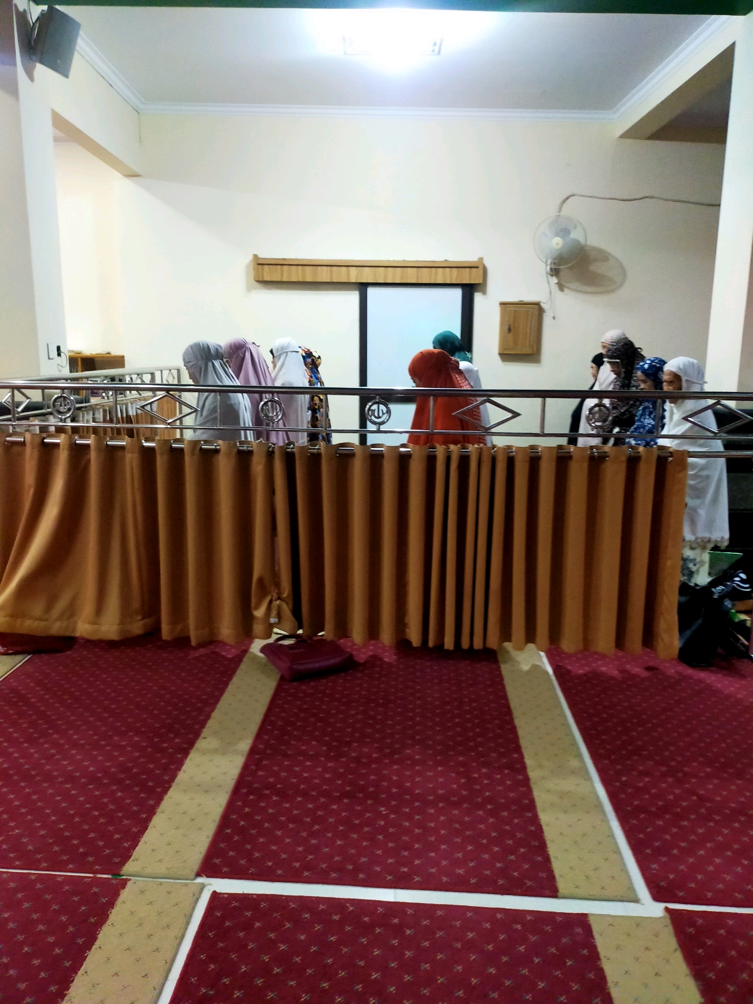 This is a medium shot of a group of women, some wearing hijabs and traditional Islamic attire, standing behind a metal railing and an orange curtain. They appear to be in a prayer hall, indicated by the red carpet with geometric patterns and the overall decor. A wall-mounted fan is visible on the right side of the frame, along with a small wooden cabinet. The lighting suggests an indoor environment, possibly a mosque. The scene is static, with no discernible action taking place. There are no landmarks or specific visual cues that would pinpoint the location to Bandung City, Indonesia.