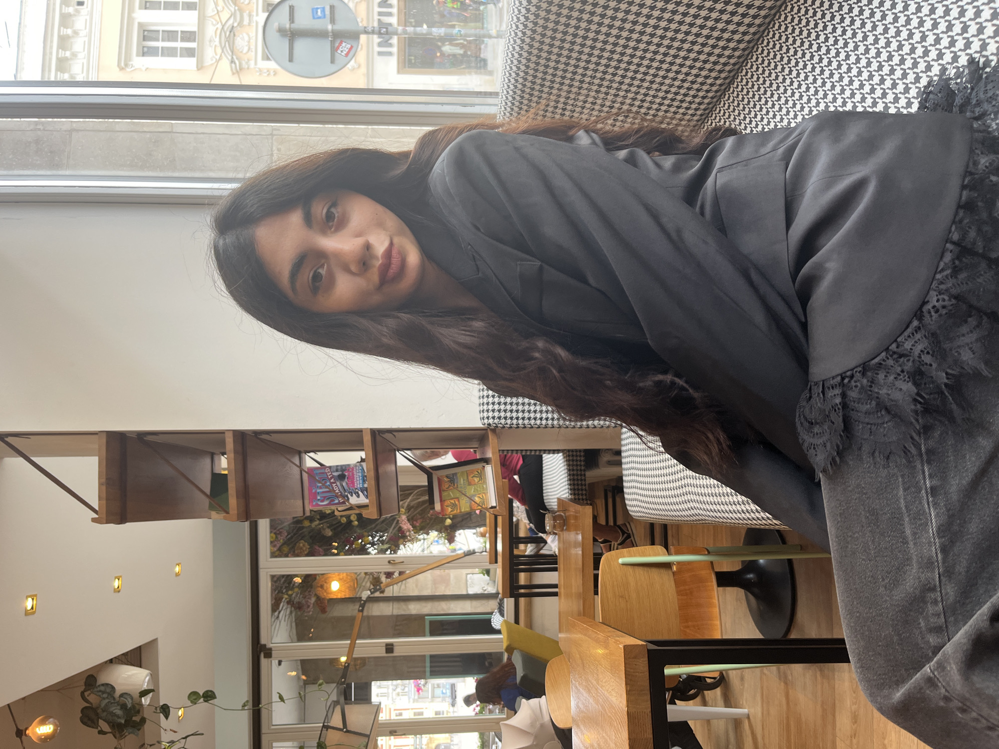 A young woman with long, dark, wavy hair is seated at a table in a cafe, looking towards the camera with a slight smile. She is wearing a black blazer over a black top and dark denim jeans that feature lace detailing at the hem. The cafe appears to be in Pilsen, Czechia, as suggested by the architectural style of the building visible through the large window behind her. The window overlooks a street with historical buildings, typical of Pilsen's city center. The interior of the cafe has wooden tables, chairs, and shelves displaying books and decorative items. A patterned bench is visible behind the woman. The overall atmosphere is casual and relaxed.