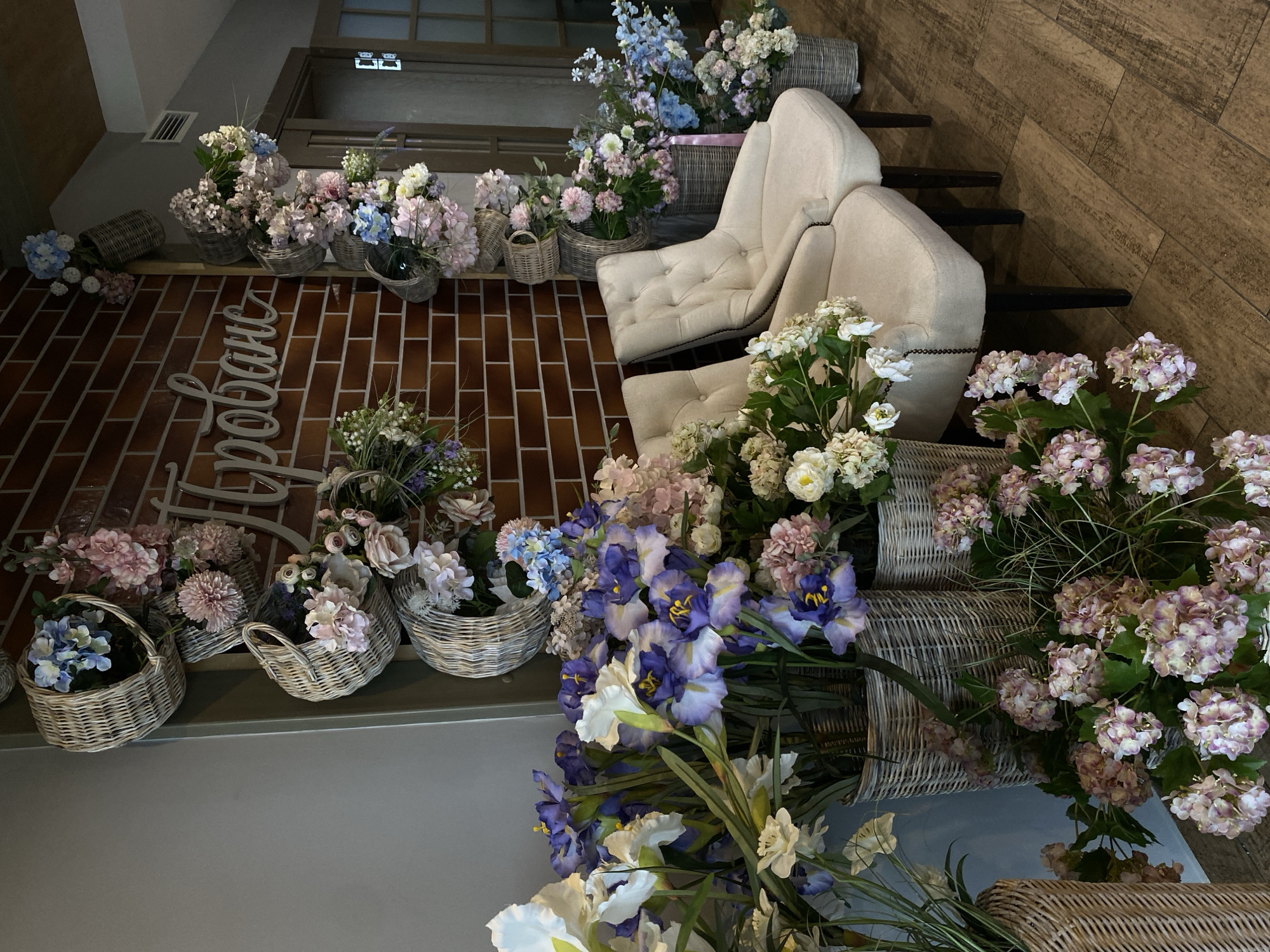 The image shows an interior space with floral arrangements and furniture. Two upholstered armchairs are visible, positioned near a staircase. The staircase has black treads and risers. The walls are covered in a brick pattern. Numerous wicker baskets filled with various flowers, including hydrangeas and irises, are arranged throughout the space. There is a decorative sign in Cyrillic script on the brick wall. The location appears to be in Votkinsk, Russia, based on the Cyrillic script of the sign on the wall. There are no people visible in the image.