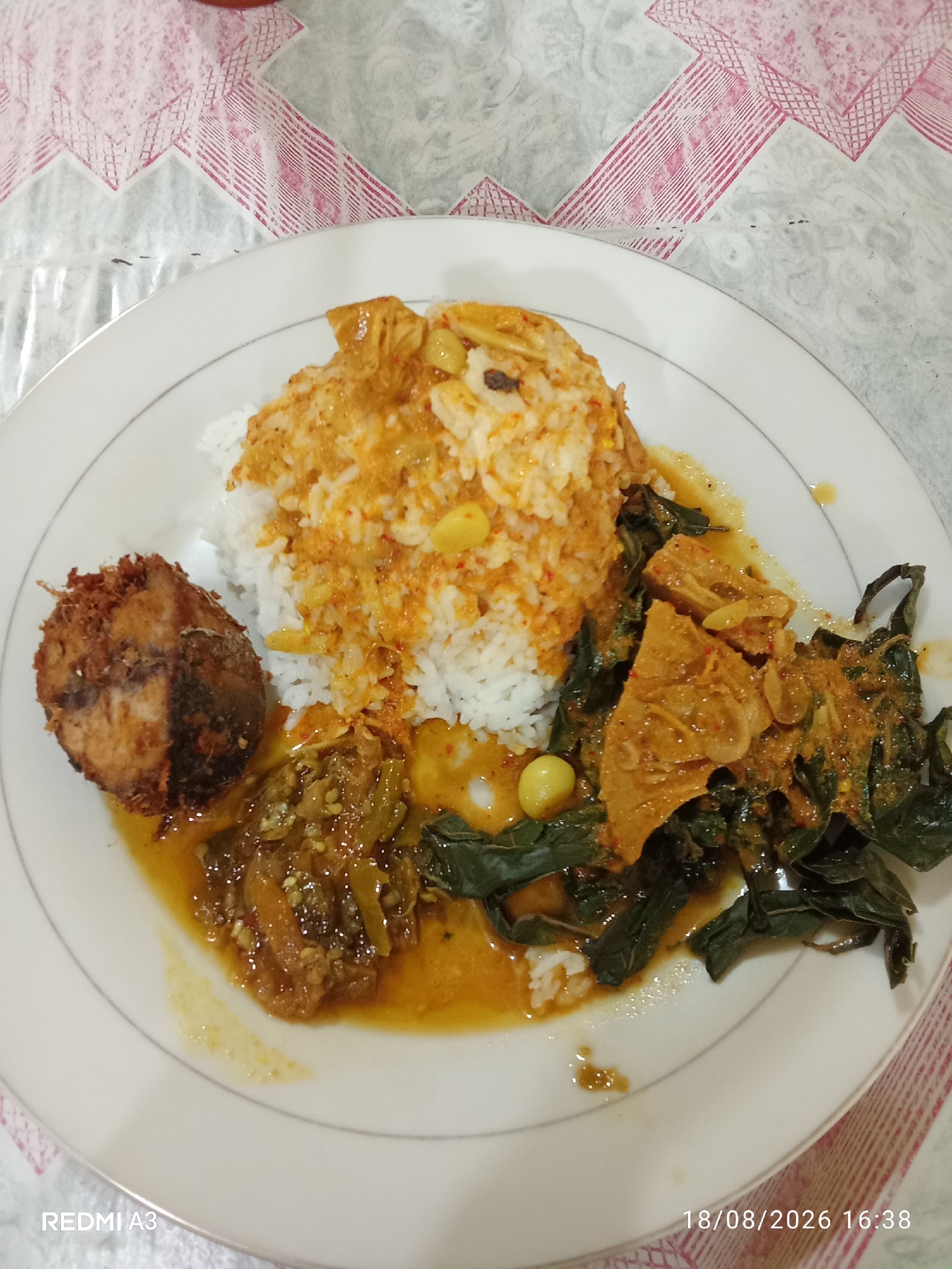 A plate of Indonesian food is pictured from a top-down perspective. The plate is white with a thin gray rim. It contains a mound of white rice topped with a rich orange curry sauce and scattered yellow pieces that appear to be beans or small fruits. To the left of the rice is a circular piece of fried food, possibly fish, with a golden-brown, crispy exterior. Below the rice and to the right is a portion of a vegetable dish, appearing to be leafy greens in a similar orange curry sauce, interspersed with chunks of what might be jackfruit or another firm ingredient. A smaller portion of a dish with a darker sauce and what seems to be meat or vegetables is located to the bottom left of the rice. The food is set on a table covered with a tablecloth featuring a geometric pattern in shades of pink and gray. The bottom left corner of the image displays the text "REDMI A3" and the bottom right corner shows "18/08/2026 16:38". The image captures a meal on a table.